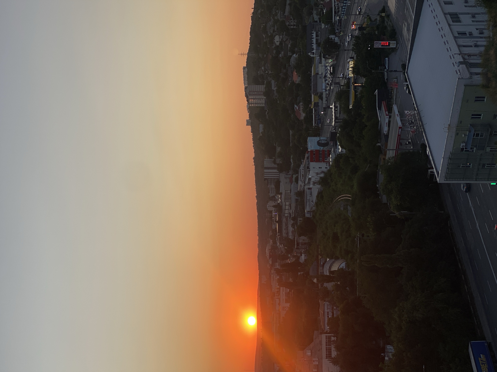 This is an aerial, vertical, wide shot of Chișinău, Moldova at sunset. The sun is a bright orange orb on the left side of the frame, setting behind a hilly landscape dotted with apartment buildings and trees. The sky transitions from a bright orange near the horizon to a pale grey at the top of the frame. Below the horizon, a city stretches out with residential and commercial buildings, roads, and a gas station with visible signage. There are no people or animals visible in the frame. The overall scene depicts an urban environment at dusk.