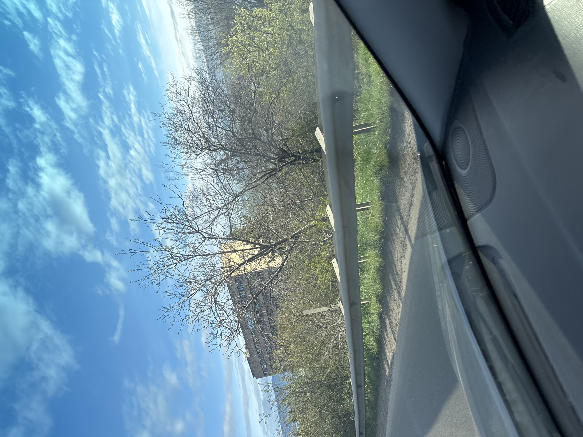 Captured through a car window, the image presents a roadside scene. To the left, a clear blue sky with scattered cirrus clouds contrasts with bare trees and budding vegetation. A multi-story building, likely an apartment complex, is situated behind the trees. A metal guardrail lines the roadside. The road surface is asphalt, with a verge of green grass. The interior of the car is visible on the right, with a partial view of the car door and speaker. Based on the architecture and the landscape, the location is potentially in the outskirts of Tbilisi, Georgia.