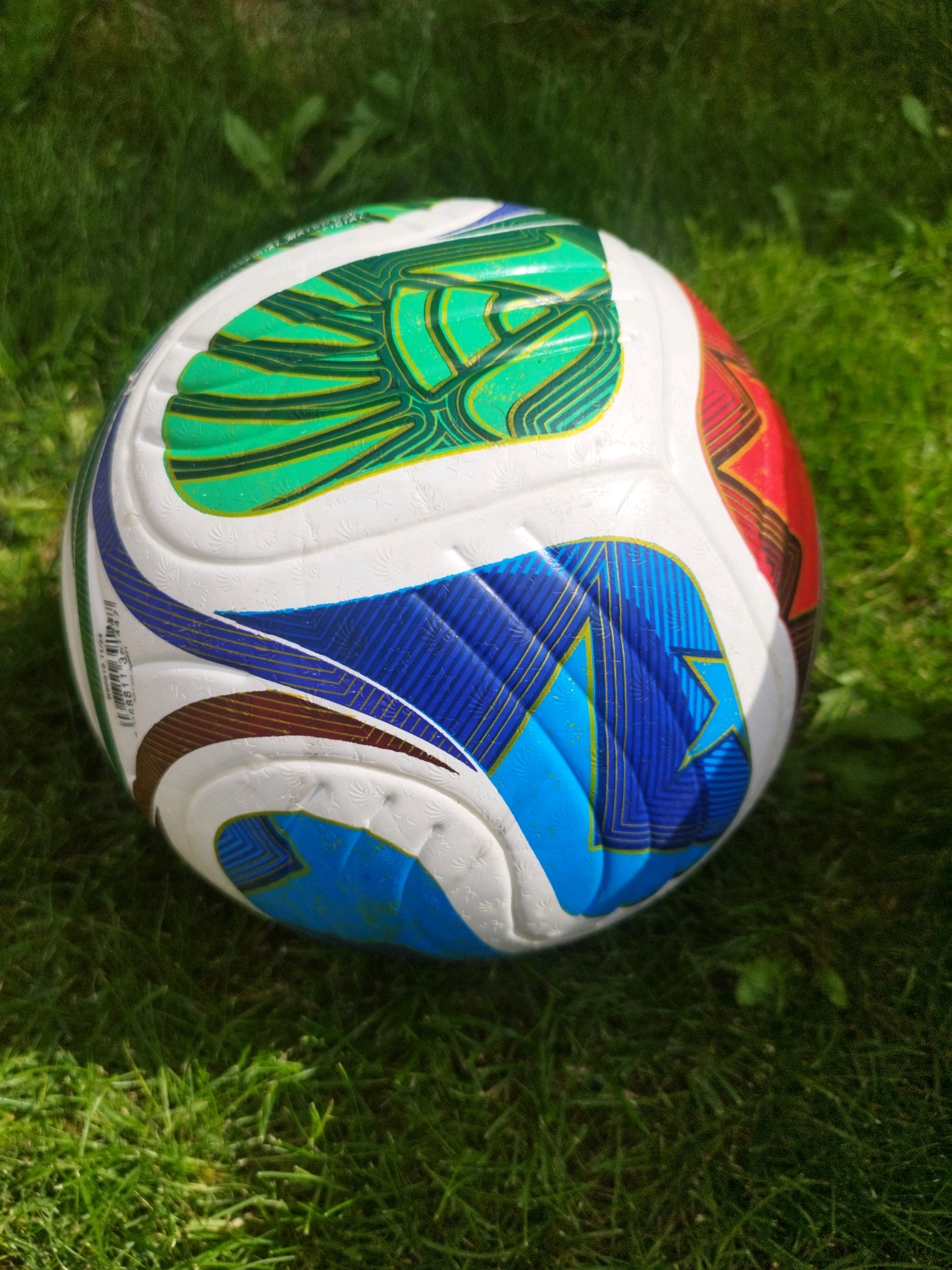 A close-up, slightly overhead shot focuses on a white and multi-colored soccer ball resting on a patch of green grass. The ball features a design with segments of green with dark green lines, blue with gold lines, and red with brown and gold lines. The grass appears well-maintained and is illuminated by sunlight, casting faint shadows. There are no people visible in the image, and no specific location in Oskemen, Kazakhstan, can be identified from the visual information provided. The scene depicts a static object in an outdoor, natural environment.