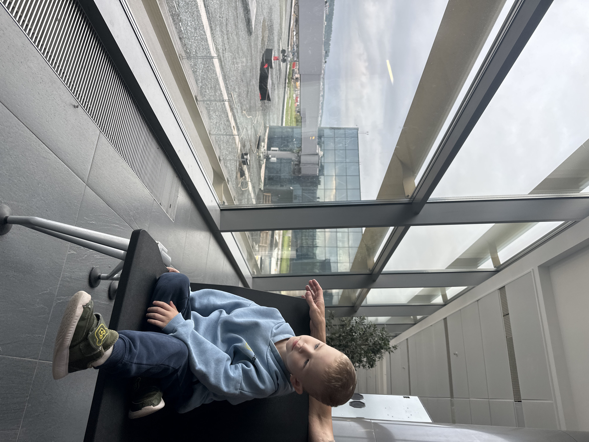 A young Caucasian boy with light brown hair and blue eyes is sitting on a black bench in what appears to be an airport or terminal building. He is wearing a light blue hooded sweatshirt and dark blue pants. His feet are resting on the floor, and he is wearing green sneakers with a teddy bear emblem. The boy is looking out a large window that overlooks an outdoor area with a jet bridge, a modern glass building, and a gray, gravel-covered ground. The sky is overcast. A person's arm is visible resting on the back of the bench behind the boy. The floor is tiled with gray square tiles, and a long, thin vent runs along the base of the window. The overall environment is modern and spacious, with large windows and metallic architectural elements. The location appears to be the airport in Bolshoye Savino, Russia, given the context provided.