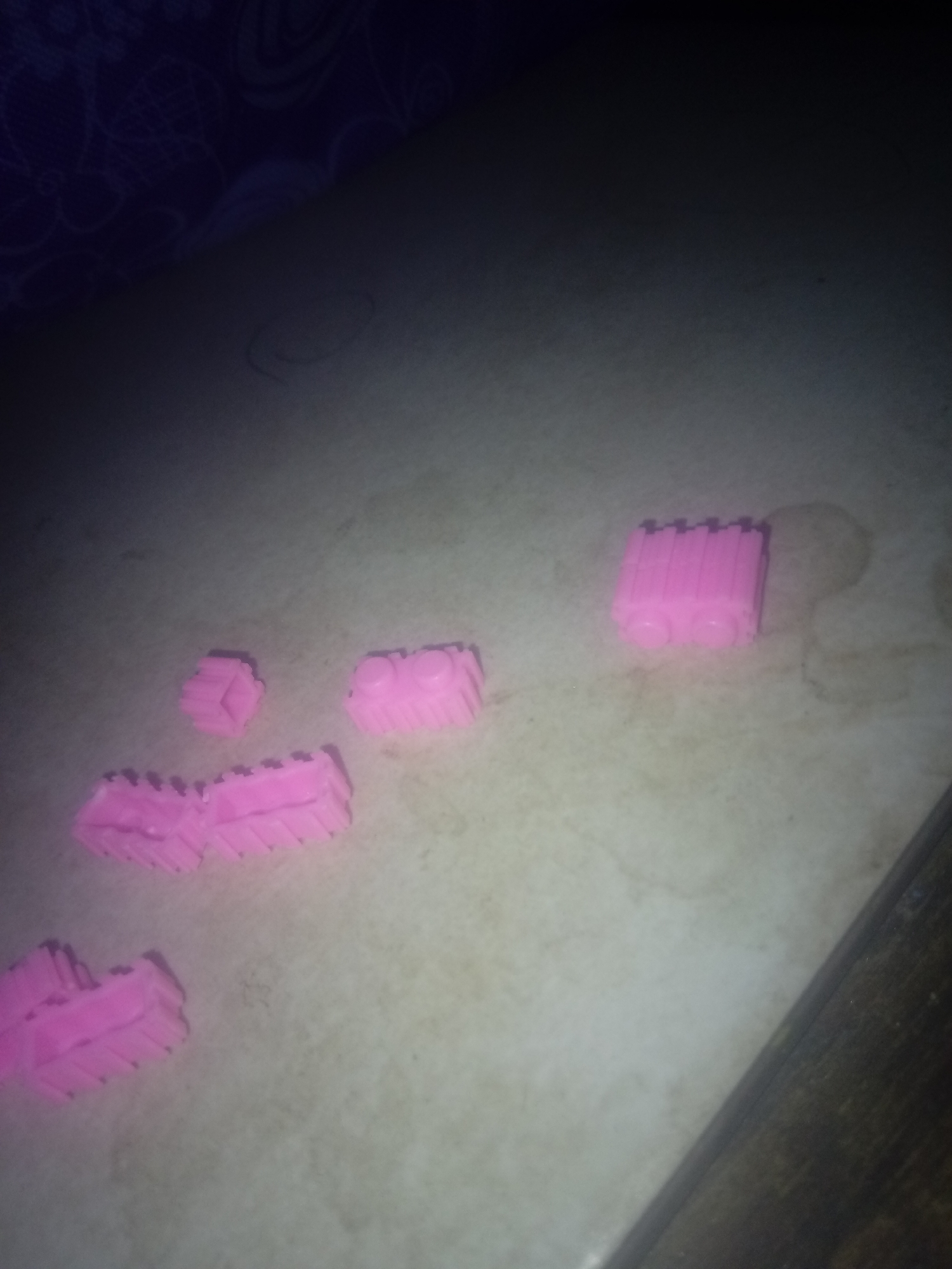 The image shows several pink toy blocks scattered on a light-colored surface. The scene appears to be indoors, with the corner of a wooden table visible in the lower right corner. The background is blurry, with a dark patterned cloth visible in the upper left corner. The lighting is dim and uneven. There is no indication that this scene is in Juan Griego, Venezuela.