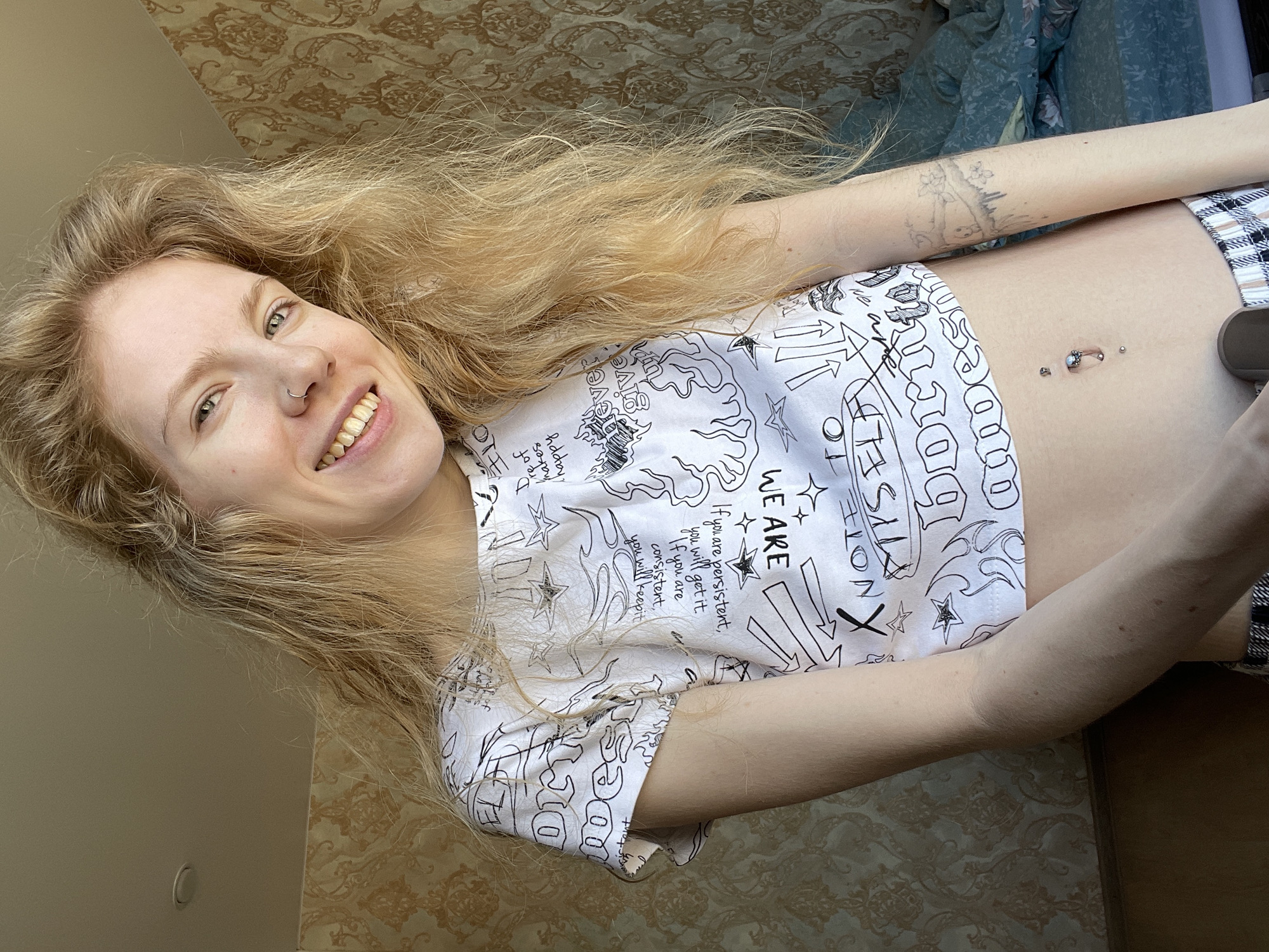 A fair-skinned young woman with long, blonde hair is smiling at the camera. She has a nose ring and visible stomach piercings. She is wearing a white cropped t-shirt with black printed designs and text. The t-shirt reads "WE ARE" and "If you are persistent, you will get it. If you are consistent, you will keep it." A tattoo is visible on her right forearm. The background includes a wall with patterned wallpaper and a partially visible blue duvet. The location (Izhevsk, Russia) is not evident from the image.