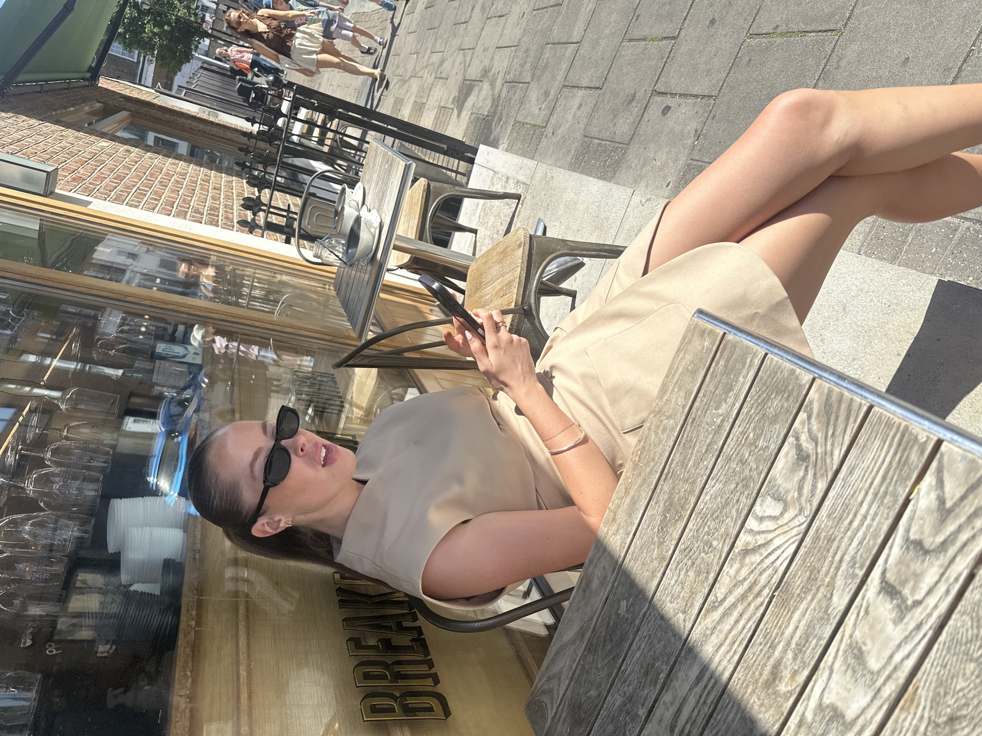 A woman with dark hair and sunglasses is sitting at an outdoor cafe in an unidentified city. She is wearing a beige dress and has her legs crossed. She is holding a smartphone. The cafe has wooden tables and chairs. The cafe window has "BREAK" visible on it. Other pedestrians are walking on the sidewalk. The location of the cafe, city, and state are all unknown.