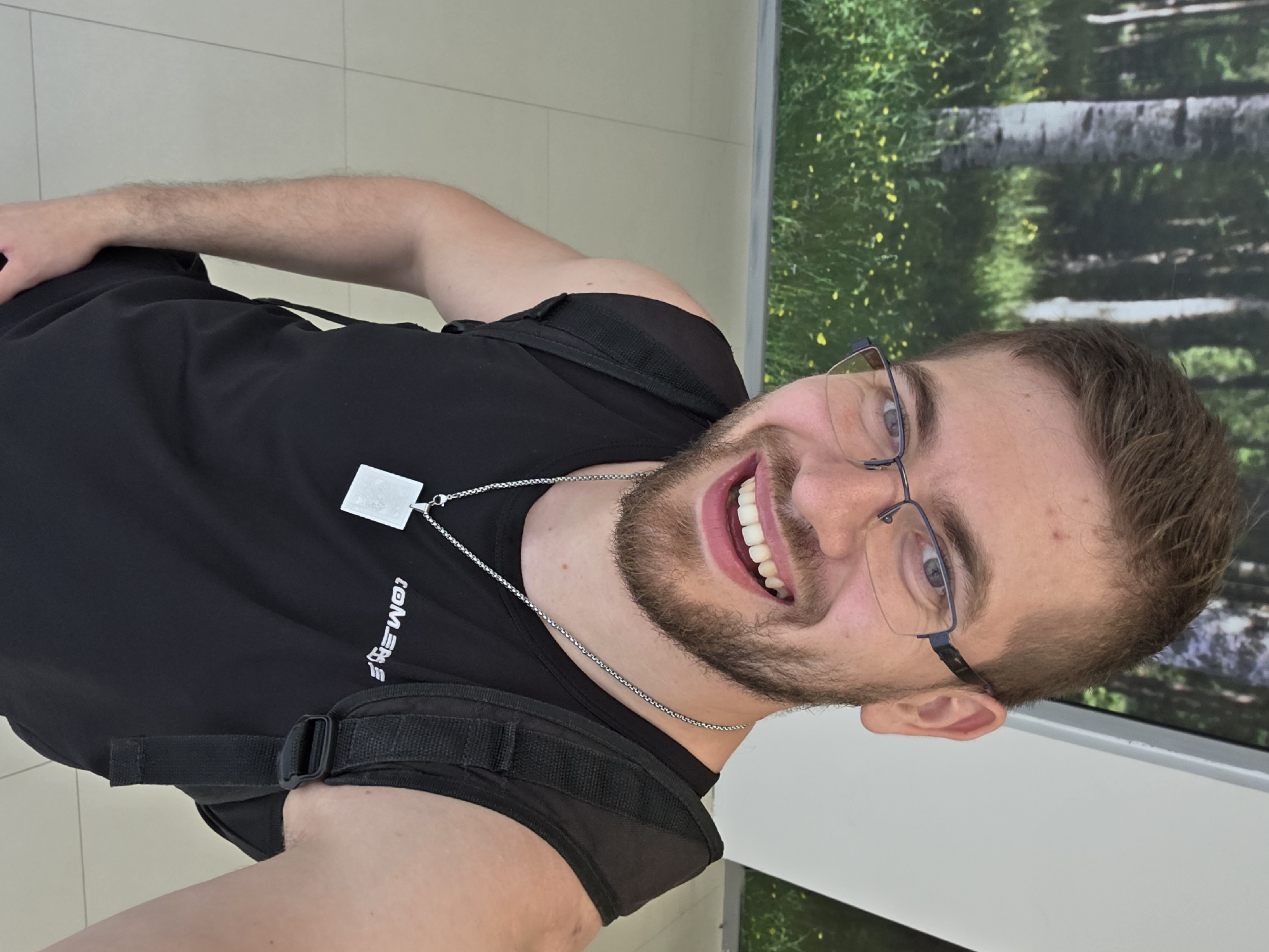 A male individual, likely in his 20s or 30s, with short brown hair and a light brown beard, is depicted in a medium-shot selfie. He wears rectangular, dark-framed glasses and smiles broadly, revealing both upper and lower teeth. He is dressed in a black sleeveless top with white text, possibly "TOMENS," printed across the chest. A silver chain necklace with a square, polished pendant hangs around his neck. A black backpack strap is visible over his left shoulder.
The background on the upper left features a large mural or poster depicting a natural forest scene with light-barked trees, green undergrowth, and numerous small yellow flowers. To the upper right, a plain white wall or pillar is visible. The floor is composed of light-colored, possibly beige, rectangular tiles with visible grout lines, indicating an indoor environment. The scene takes place within an unidentified indoor public space in the city of Plovdiv, Bulgaria.