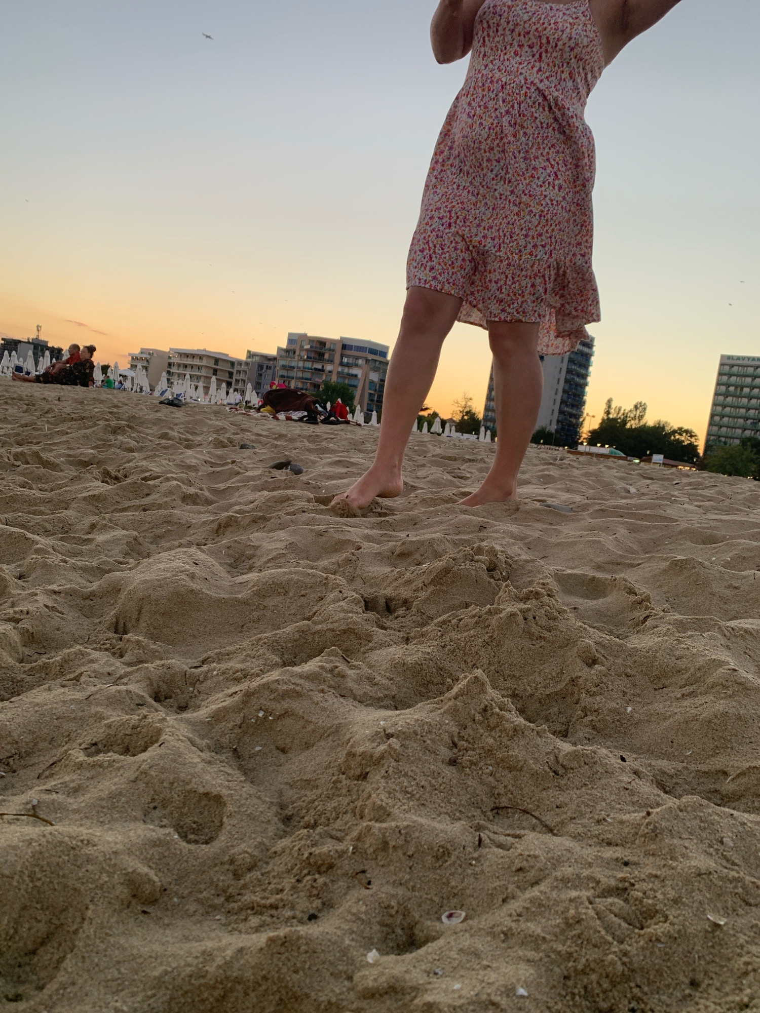 The image captures a sandy beach in Nesebar, Bulgaria, during what appears to be sunset or sunrise. In the foreground, the focus is on the rippled texture of the sand, with some small shells and debris visible. In the midground, the legs and lower torso of a person wearing a floral dress are visible, standing on the sand. Their feet are bare. To the left and further in the background, several other people are seated on the sand, and beach umbrellas are set up. The background features a line of modern, multi-story buildings and trees against a pale orange and light blue sky. A bird is visible in the sky. The scene suggests a peaceful, recreational setting.