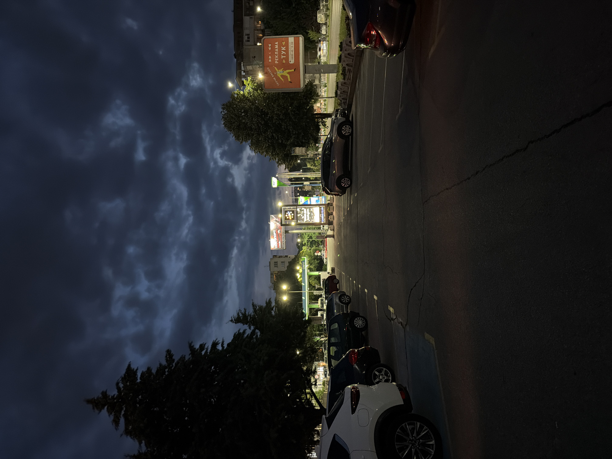 The image depicts a nighttime scene in a parking lot in Plovdiv, Bulgaria. The sky is dark with heavy, dramatic clouds. In the foreground, the asphalt parking lot is visible with white parking lines. Several cars are parked in the lot, including a white SUV on the left, a dark SUV, and other dark-colored vehicles.
In the background, there are trees, streetlights, and several billboards. One prominent billboard, with an orange background and a depiction of a running figure, displays Cyrillic text including "РЕКЛАМА" (REKLAMA - advertisement) and a phone number. Another billboard features bright lights and Cyrillic text that appears to be for a "CAMPING CLUB". A gas station with an OMV sign is also visible. The overall atmosphere is slightly overcast and dim, typical of an evening in an urban environment. No people are visible in the image.