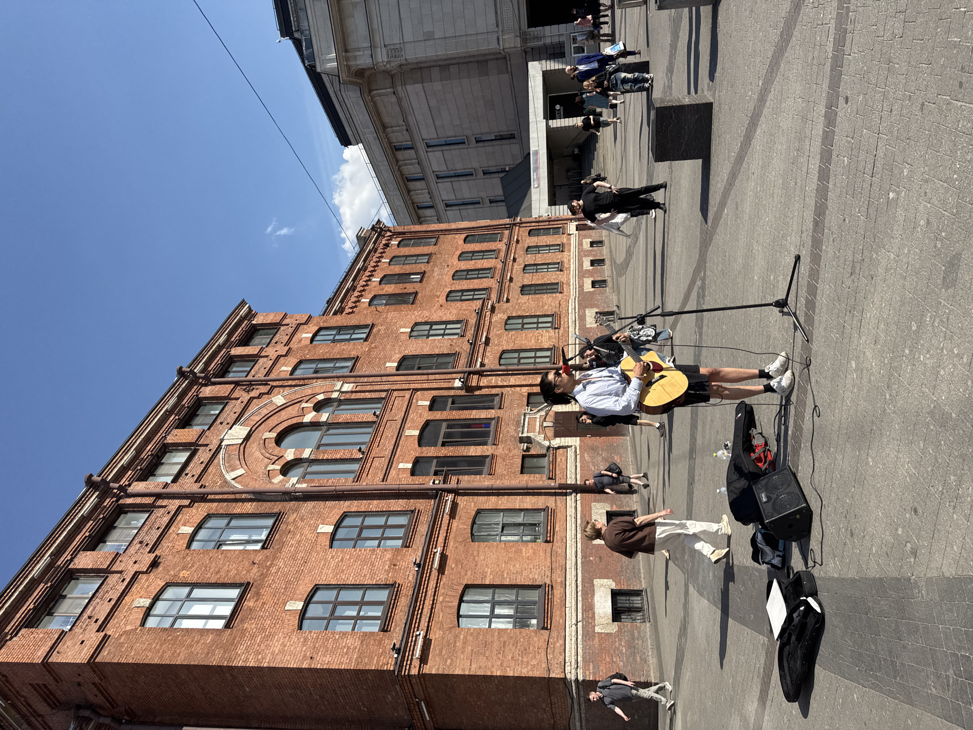In Saint Petersburg, Russia, on a paved square in front of a large, red brick building, a street performer is playing an acoustic guitar. The performer, wearing a light-colored collared shirt, dark shorts, and white sneakers, stands with a microphone in front of them and their guitar. Their equipment, including a black amplifier, a guitar case, and a small white object, is laid out on the ground nearby.
Several other people are walking around the square. Some are walking towards the performer, while others are moving away or passing by. The backdrop features a tall, light-colored building with a modern architectural style. The sky is clear and blue with a few wispy clouds. The sun is shining, casting shadows on the pavement. The area appears to be an open public space with a mix of historical and contemporary architecture.