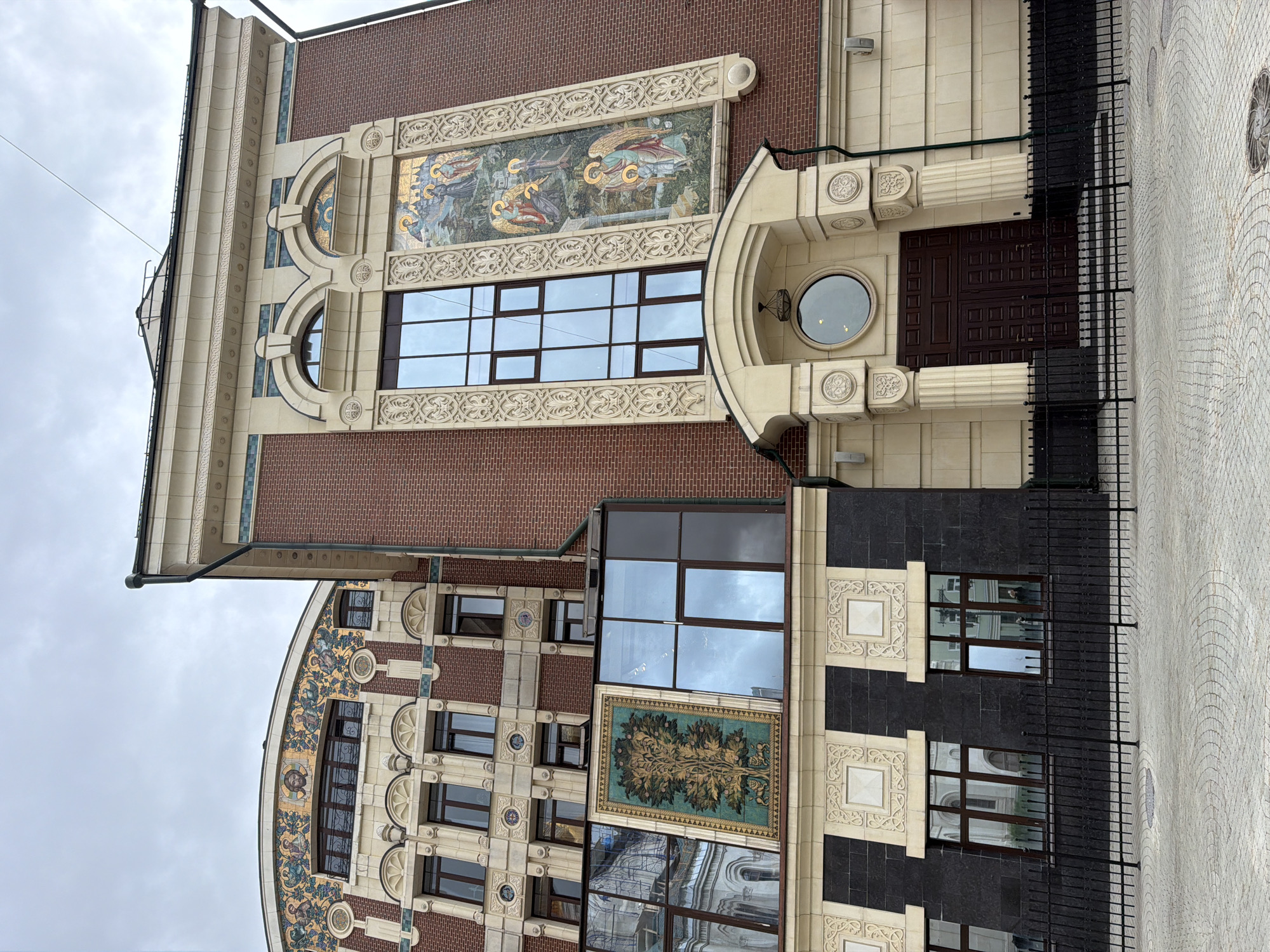 The image shows the facade of a building with distinct architectural features. The building is primarily constructed of red brick, with large sections of light-colored stone and decorative mosaics. One mosaic panel, located on the right side of the building, depicts religious figures, possibly angels and a child, in a landscape. Another mosaic panel on the left side of the building features a stylized tree with animals at its base.
The building has numerous windows, some of which are large and modern-looking with dark frames, while others appear older and more traditional. The entrance features a large, dark wooden double door beneath a rounded stone archway. A black metal fence runs along the front of the property, and the ground in front of the building is paved with light-colored cobblestones. The sky is overcast.
This building is the Moscow Choral Synagogue, located at Bolshaya Nikitskaya Street, 42, in Moscow, Russia.
