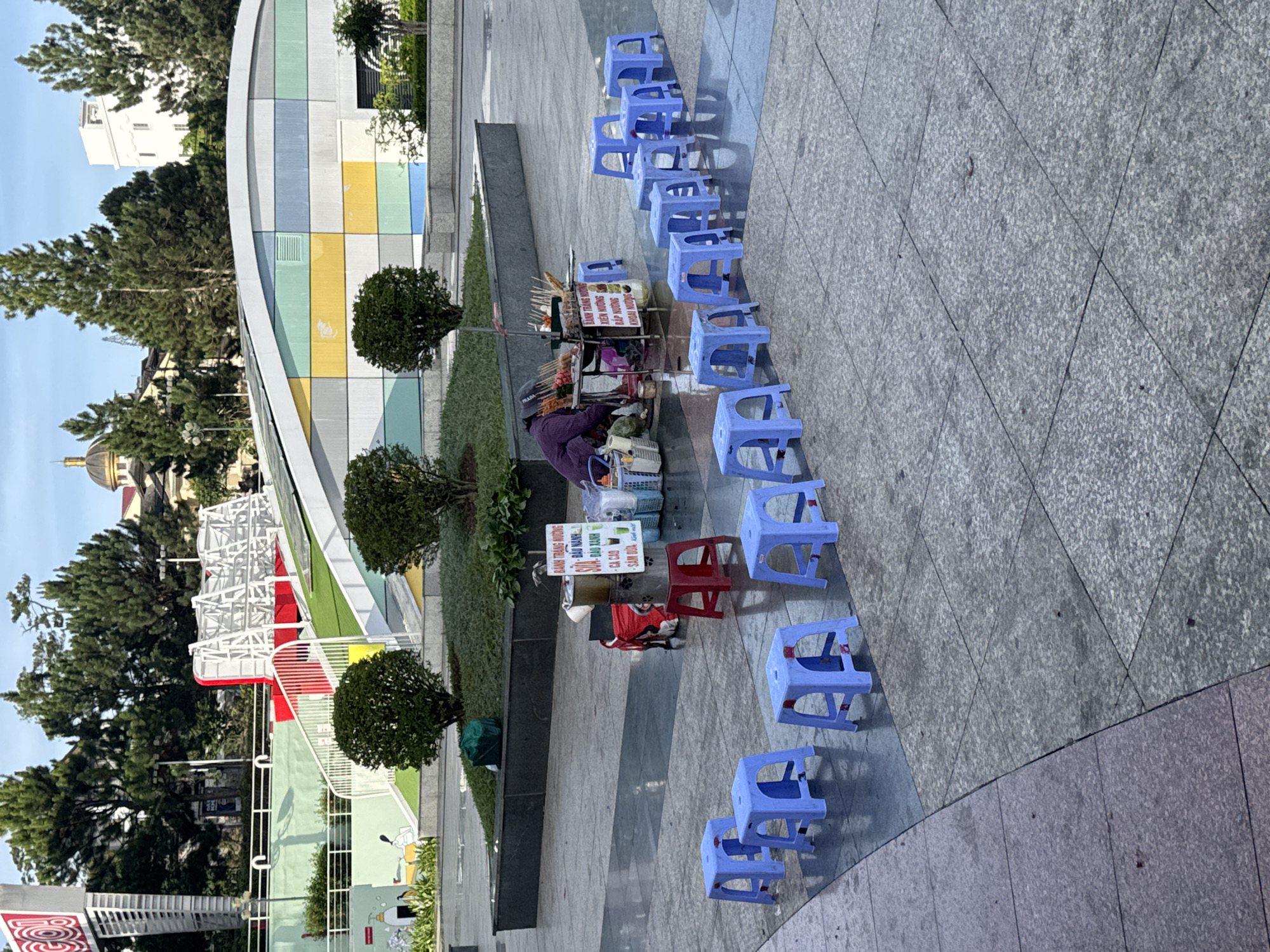 A vendor in Da Lat, Vietnam, sells street food from a cart along a modern walkway with blue plastic stools arrayed for customers. A woman in a purple shirt attends the cart, which displays food on sticks and a sign in Vietnamese. A couple of red stools are present among the blue ones. The walkway consists of gray concrete tiles with alternating textures. Behind the vendor are manicured hedges and a colorful building facade. In the background, trees obscure other buildings, including one with a dome.