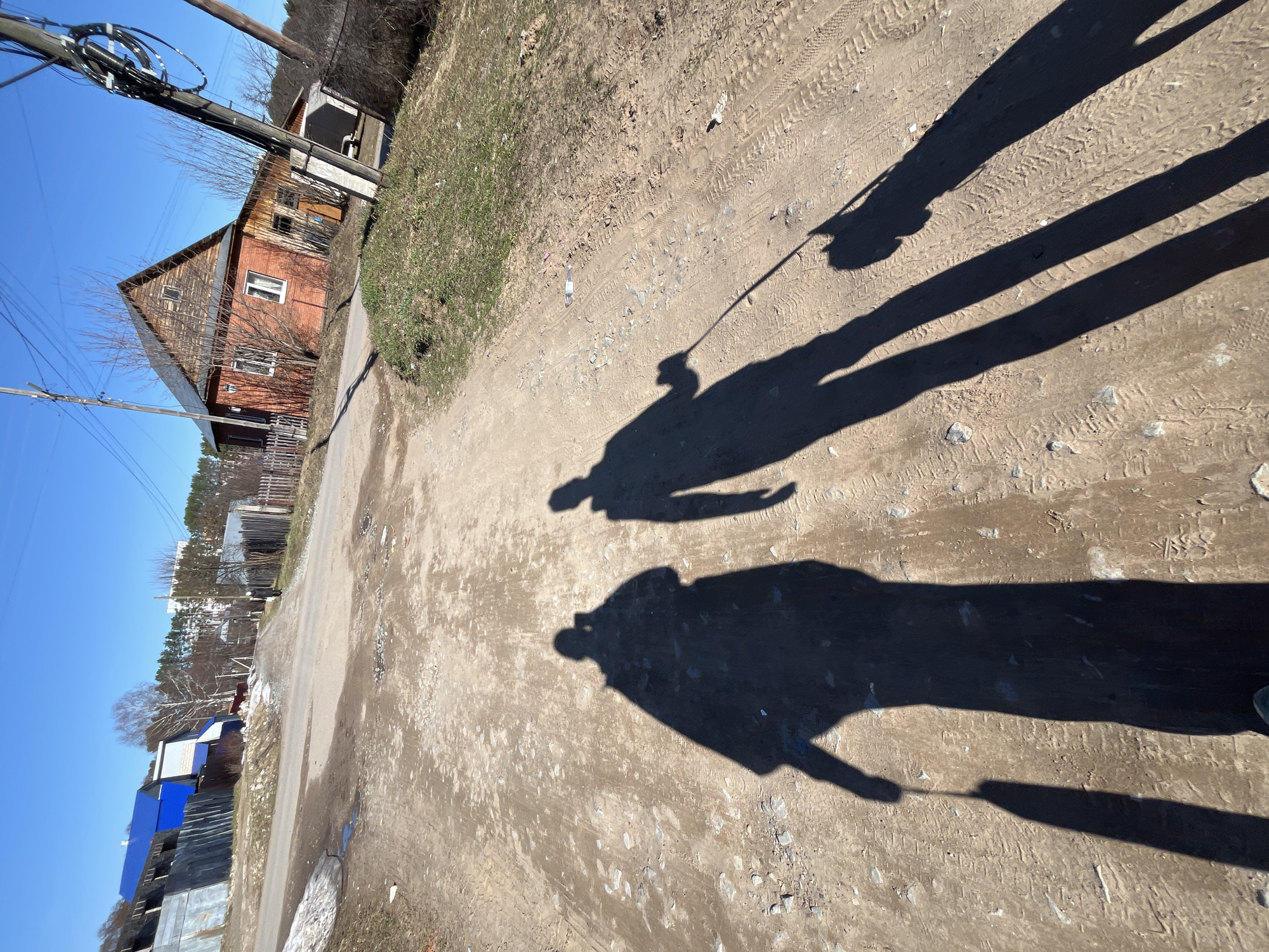 The image depicts a dirt road in what appears to be a rural or semi-rural area. Three shadows are cast onto the road, suggesting the presence of three individuals. The shadow furthest to the right has a visible arm holding some kind of stick. Buildings, including a two-story wooden house with a gabled roof, are visible to the left of the road. Utility poles and wires are also present. The sky is clear and blue.
Based on the description, the image most likely reflects the area of Chaikovsky, Russia. While I cannot provide an exact location without additional information, the scene is consistent with the more rural or residential neighborhoods of that city.