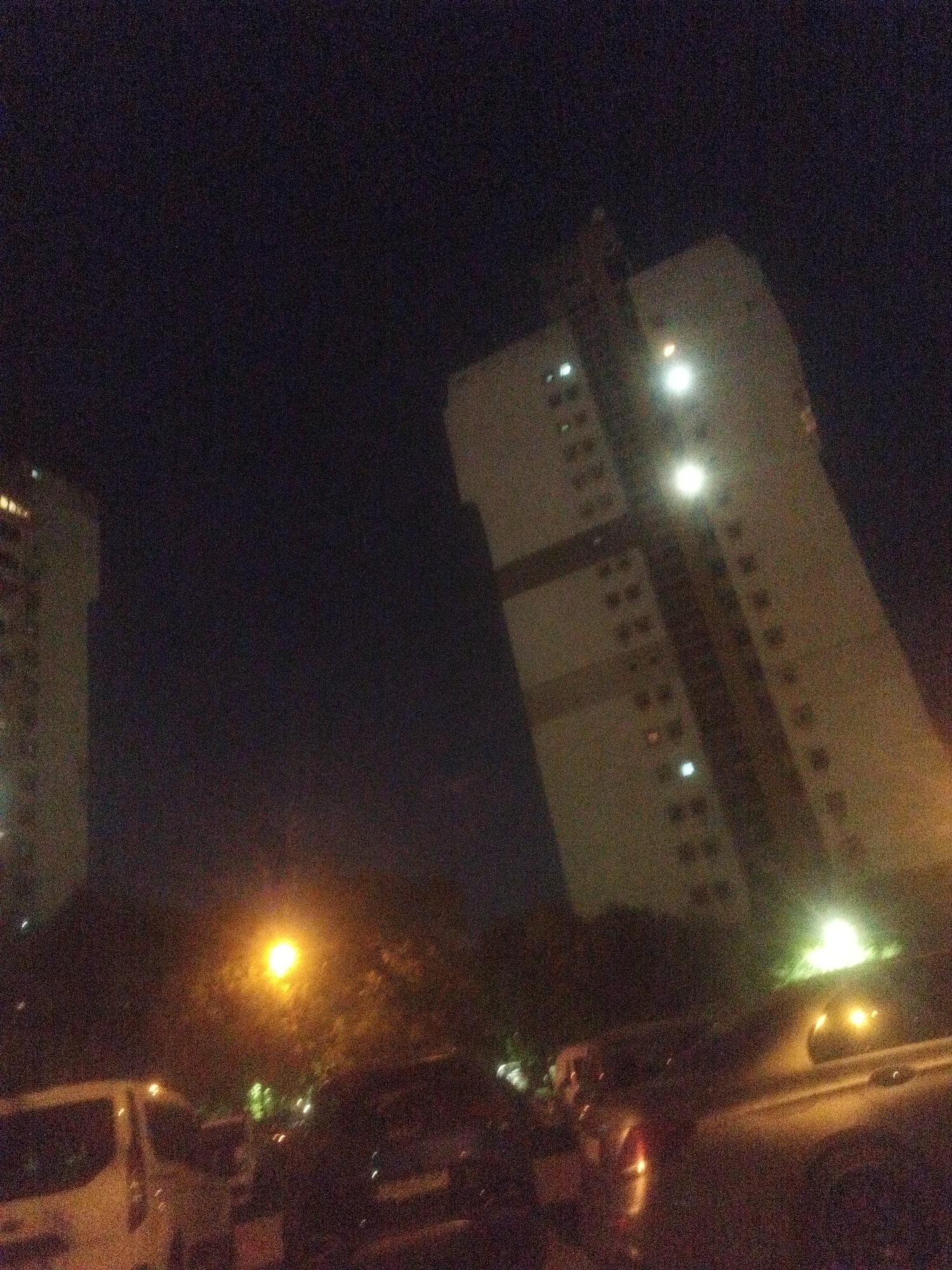 The image shows a nighttime scene, likely in an urban area based on the visible structures. Two tall buildings with multiple floors are visible, illuminated by interior lights. Between the buildings and the camera, there's a row of trees and streetlights, and a line of parked cars in the foreground. The overall scene suggests a residential area or a mixed-use zone. The image quality is relatively low, with significant noise. Based on the architecture and street layout, it's possible that the image was taken in Sofia, Bulgaria.