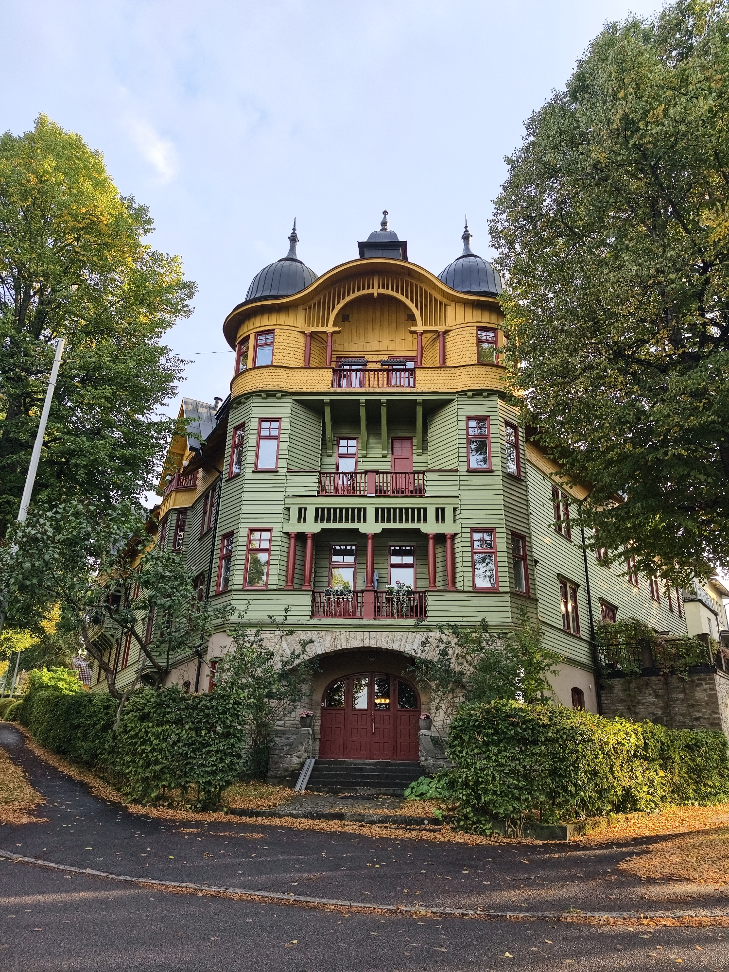 The image depicts a tall, historic building with a distinctive green and yellow facade, featuring multiple balconies and decorative elements. The building is situated on a slightly elevated area, with a winding road and fallen autumn leaves in the foreground. Two large, mature trees frame the building on either side, their leaves showing signs of autumn coloration. The sky above is a pale blue with a few wispy clouds. The architectural style suggests it might be from the late 19th or early 20th century. The location is identified as the Singer building at Ringgatan 26 in Norrköping, Sweden. The building has a prominent rounded corner with a domed roof section, and a grand arched entryway with red double doors. The overall impression is one of an old, well-maintained, and visually striking structure in a natural setting. There are no people visible in the image.