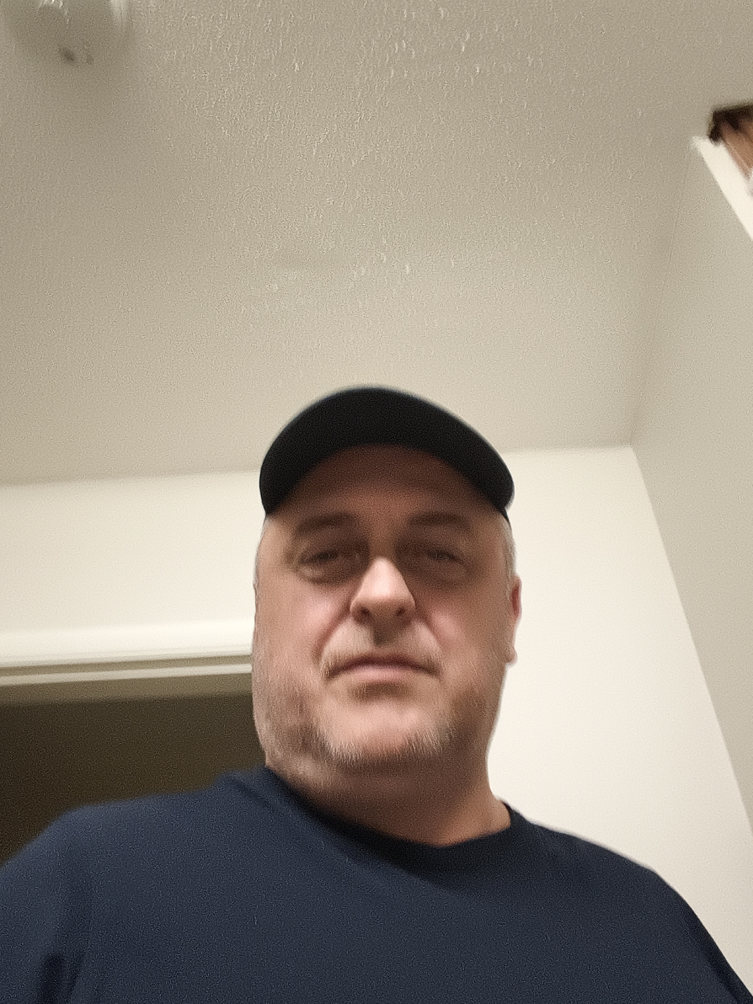 The image features a man taking a selfie or being photographed indoors in Huntingdonshire, United Kingdom.
The subject is a middle-aged man with fair skin and light facial hair, appearing as stubble or a short beard. He is wearing a dark-colored baseball cap, positioned with the brim forward, and a plain, dark blue short-sleeved t-shirt. His expression is neutral as he looks directly into the camera.
The photograph is taken from a slightly low angle, looking upwards at the man. The background consists of plain, light-colored walls and a ceiling, possibly off-white or light beige. The ceiling and walls exhibit a textured, somewhat grainy appearance, which is more clearly visible in the provided crops.
On the left side of the frame, a dark-colored door frame is partially visible in the blurred background. In the upper left corner of the ceiling, there's a faint, dark, indistinct object, possibly a light fixture or a mark. A corner where two walls meet is visible on the right side of the background.
The lighting appears to be artificial and internal, consistent with an indoor setting, and does not provide cues for the time of day or outdoor weather. The overall image quality shows some digital noise, particularly noticeable on the plain wall and ceiling surfaces.
There is no visible text in the image.