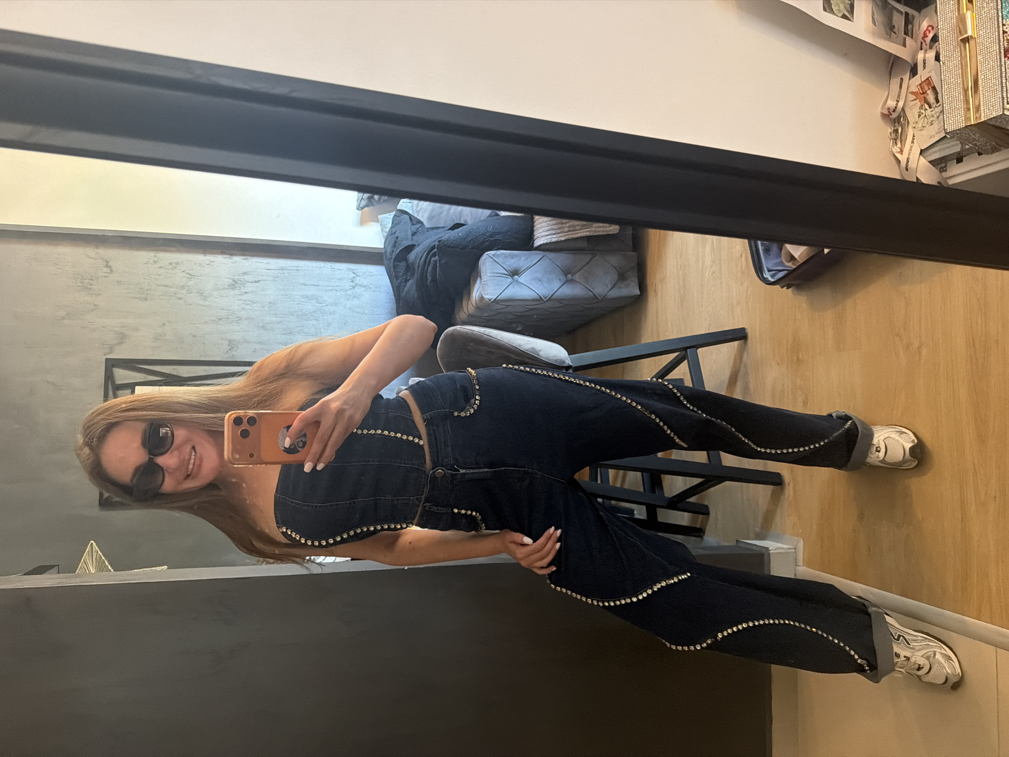 A young woman is captured in a mirror selfie, showcasing a stylish dark denim two-piece outfit heavily embellished with small, shiny studs or rhinestones. She stands at a slight angle, smiling and wearing dark sunglasses. Her long, light brown hair falls over her shoulders, and her left hand rests on her hip or upper thigh, while her right hand holds an orange-brown smartphone to take the picture. She has white painted nails and rings on her fingers.
Her outfit consists of a strapless or corset-style top and matching wide-leg or flared jeans. Both pieces feature wavy patterns of the embellishments. The jeans are cuffed at the bottom, revealing white athletic sneakers with gray accents.
The background, reflected in the mirror, shows an indoor setting. To the left, there's a light-colored wall transitioning to a textured dark gray wall. A dark gray or black plush, tufted armchair or ottoman with blankets and pillows is visible behind her. The floor is light wood. To the right, shelves or display units hold various items, including what appear to be lanyards or ribbons with some text, possibly "DREAM GROUP," which is largely illegible due to the reflection and distance. A dark metal furniture frame is also visible on the floor near her legs. The overall ambiance suggests a modern and well-appointed interior, possibly a boutique, dressing room, or stylish home in Bar, Montenegro.
