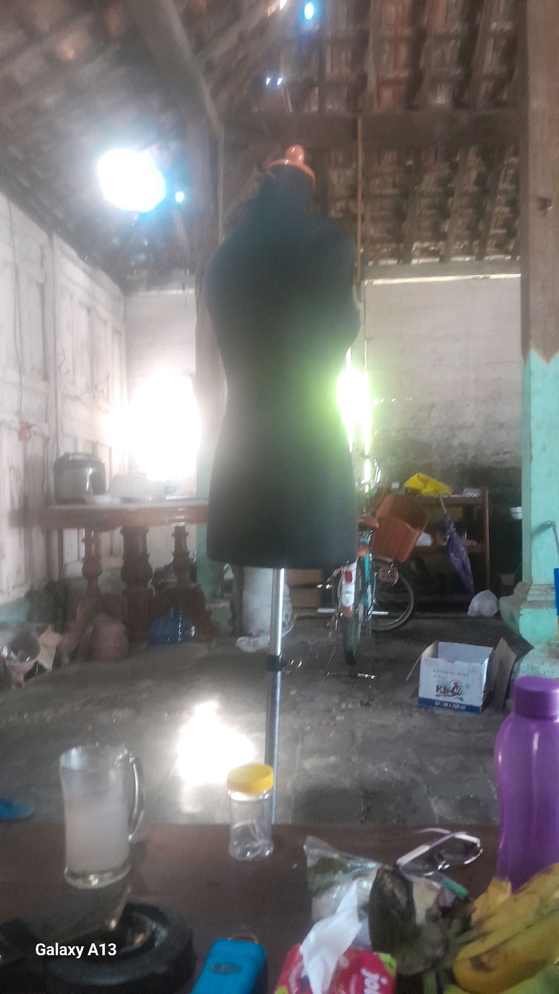 In Ngelo, Indonesia, an indoor scene unfolds within what appears to be a rustic workshop or storage area. A black mannequin, standing upright on a metal pole, is the central focus. To its left, a wooden table is laden with various objects, including a black appliance, possibly a pot, and a white bowl or container. Behind the table, a white wall with vertical paneling is visible. Further into the background, to the right of the mannequin, a bicycle with a basket on the front is parked. Shelving units filled with items and a purple bottle stand against the wall. The floor is concrete, scattered with debris, and a cardboard box is placed near the foreground. A glass of light-colored liquid and a small transparent container with a yellow lid sit on a surface in the lower foreground. The overall lighting suggests daylight filtering in, creating bright spots and casting shadows, indicating a somewhat dim interior. The environment feels utilitarian and somewhat cluttered, characteristic of a working space.