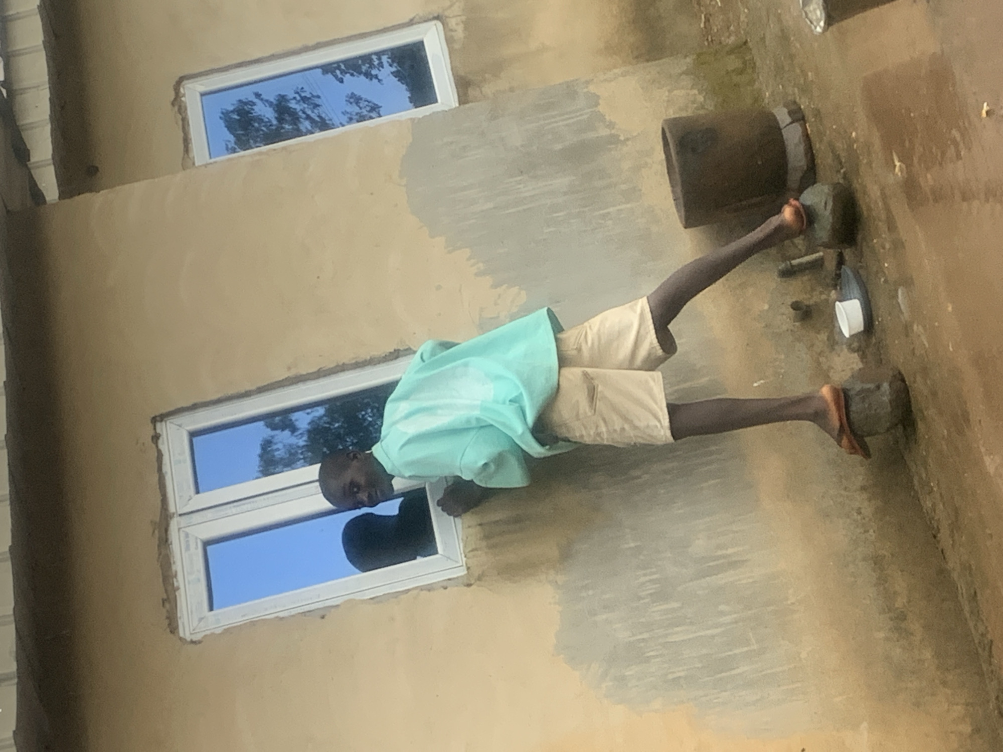 In Agban, Nigeria, a young boy stands on two stones outside a building. He is wearing light-colored shorts and a blue-green shirt. The building has two windows with blue reflective glass and a textured, light brown wall. One window is larger and features a reflection of the boy and another person. The ground is uneven and appears wet in places. A dark, cylindrical object resembling a mortar is visible to the boy's right, along with a small white container on a plate to his left. The scene depicts an outdoor setting near a residential structure.