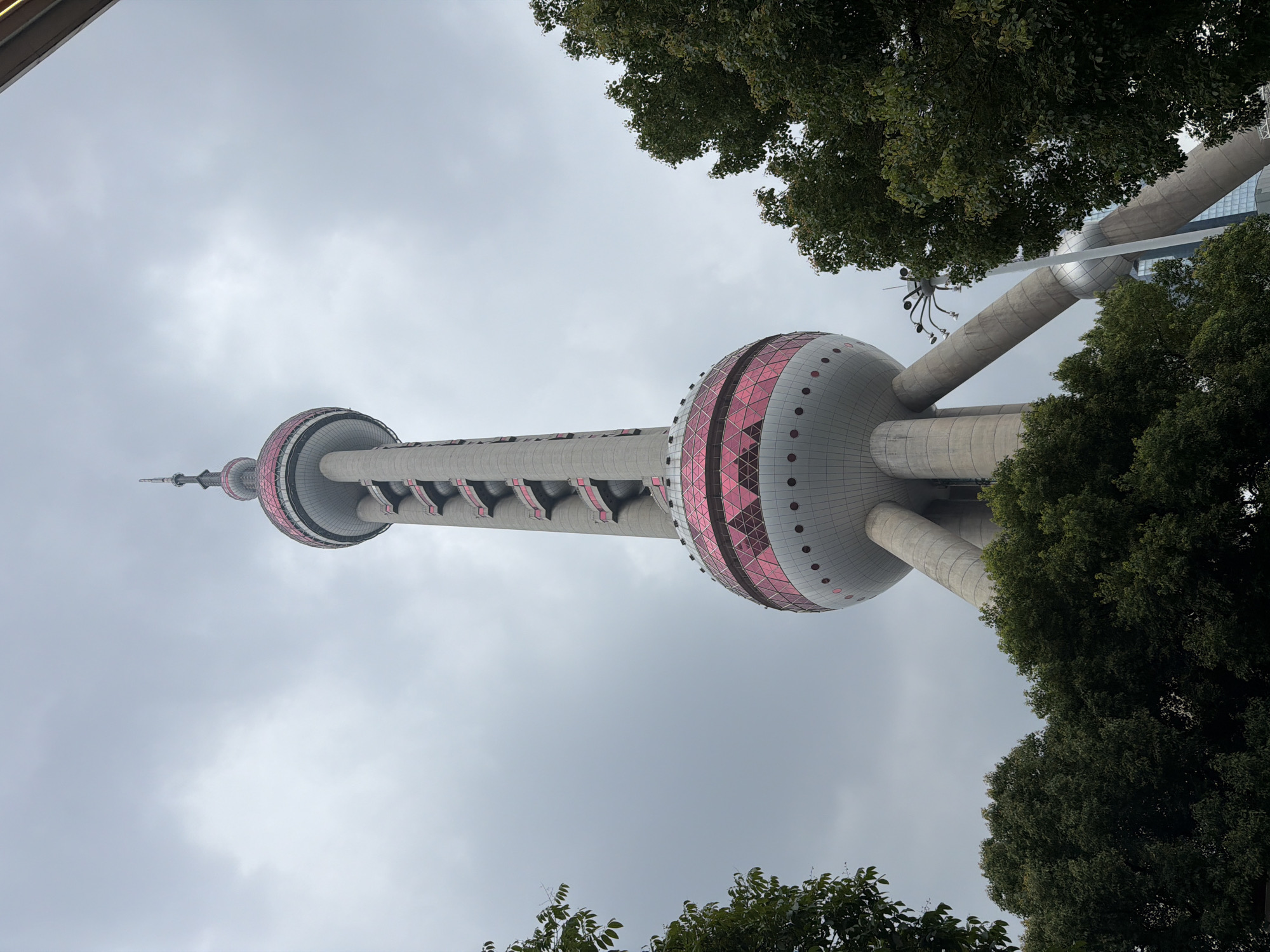 A low-angle, full-shot photograph captures the Oriental Pearl Tower in Pudong, China, against a cloudy sky. The tower, a prominent landmark, dominates the frame with its distinctive design. It features a slender, cylindrical body with three large spheres, the upper two adorned with a pink, geometric pattern and the lower one exhibiting circular cutouts. The tower is supported by four massive concrete legs that spread outwards. Green trees partially obscure the base of the tower and frame the bottom of the image. A modern building and a streetlamp are visible in the background, peeking through the foliage. The overall lighting suggests an overcast day, contributing to a somewhat muted color palette.