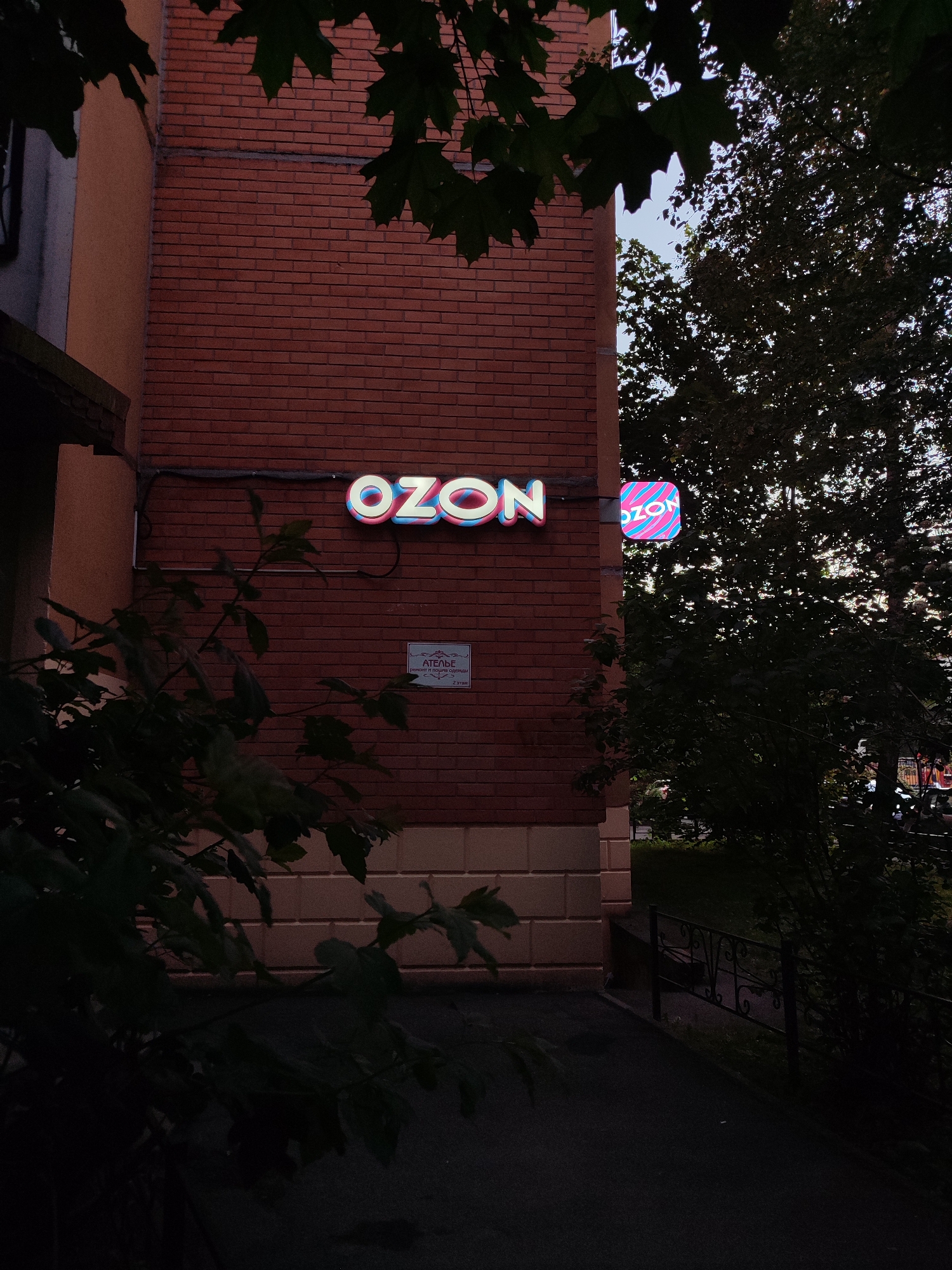The image captures an exterior view of a brick building in Vsevolozhsk, Russia, featuring a prominent illuminated sign for "OZON." The sign's letters, "OZON," are rendered in a neon style with alternating pink and blue colors. To the right of this sign, a smaller square sign also displays the "OZON" logo, which consists of a stylized pink and blue striped design. Below the larger "OZON" sign, a smaller, rectangular sign with text in Cyrillic indicates "АТЕЛЬЕ" (Atelier) and "ремонт и пошив одежды" (clothing repair and tailoring), with "2 ЭТАЖ" (2nd floor) written below. The building's facade is constructed from reddish-brown bricks and has a beige-colored lower section with decorative horizontal lines. Lush green foliage from trees and branches partially obscures the building and signage, suggesting an outdoor setting with natural elements present. In the background, a black metal fence and greenery are visible, hinting at a walkway or path leading away from the building. The overall lighting suggests it is either dusk or an overcast day, with the neon signs providing points of illumination.