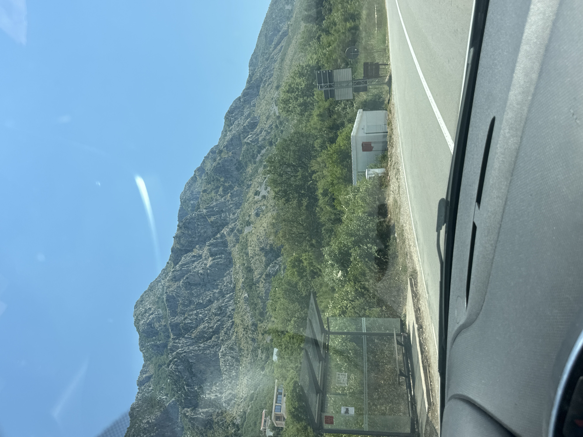 The image captures a view from a vehicle dashboard, looking out towards a rural road scene in Blizikuće, Montenegro. The foreground shows the dark grey dashboard and windshield. In the mid-ground, a paved road runs horizontally across the frame, with a white lane marker. To the left of the road is a bus stop shelter with a transparent roof and glass sides, containing a wooden bench. A red sign is visible on the side of the shelter. Adjacent to the bus stop is a small, white, box-like structure with a red mailbox on its side. Further to the right, a metal pole with signage stands near the road. Behind these elements, a dense green forest covers a sloping hillside. Dominating the background is a large, rugged, grey mountain range, with its rocky slopes partially covered by vegetation. The sky is a clear, bright blue with a faint, horizontal streak of light across it. A few modern buildings with large windows are visible on the upper part of the hillside, nestled among the trees. The overall impression is a sunny day in a mountainous, coastal region.