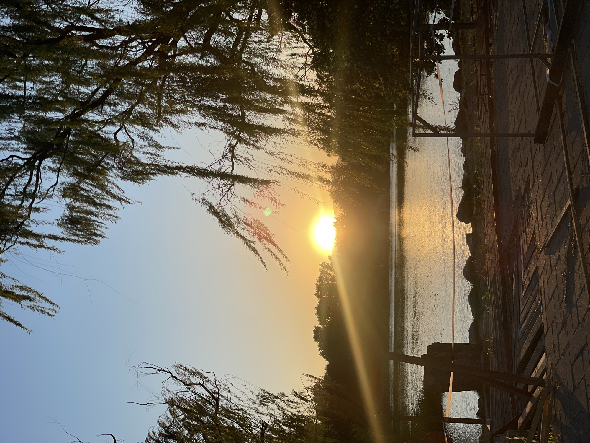 The image depicts a sunset over a body of water, possibly a lake or river, with trees framing the scene. The sun is partially obscured by foliage, casting a golden glow across the water. On the right side of the image, there is a structure that appears to be a boardwalk or path with a railing. The location is Haidian District, China.