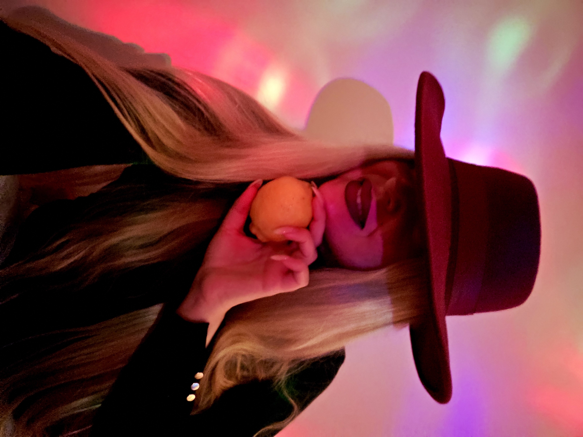 An upward-oriented, eye-level shot captures a person with long, flowing blonde hair wearing a dark, wide-brimmed hat and a dark garment, possibly a jacket, which has three small buttons visible on the sleeve. The person holds a bright yellow citrus fruit, resembling a lemon or small orange, in their left hand, bringing it close to their mouth. Their face is largely obscured by the hat and hair, though red lips are visible beneath the fruit, possibly highlighted by makeup or ambient light.
The scene is dominated by a vibrant, multi-colored background, featuring diffused light in shades of pink, purple, red, and hints of green and yellow. This colorful wash suggests an indoor setting with dynamic, artificial lighting, perhaps a club, concert, or artistic performance space. The lighting creates a dreamy, somewhat abstract atmosphere. The person's pose is stylized, seemingly mid-action or a deliberate artistic composition. No text is visible in the image.