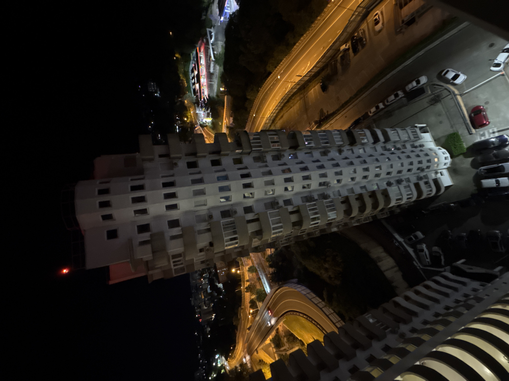 The image is a nighttime aerial view of a city, likely Sochi, Russia, featuring a distinctive, uniquely shaped building. The building, with its multi-faceted, almost crystalline facade and numerous balconies, dominates the foreground. A red warning light is visible at the very top of the structure. Below and to the left, a multi-lane highway curves through the urban landscape, illuminated by streetlights and the headlights of a few cars. The highway appears to be elevated in sections, with supporting structures visible. To the right of the building, another section of highway is visible, also well-lit. In the background, a city skyline with scattered lights stretches into the darkness, suggesting a moderately populated urban area. Further in the background, what appear to be gas stations with illuminated roofs are visible. The overall impression is that of a modern, somewhat geometrically striking architectural feature situated within a bustling, illuminated urban environment at night.