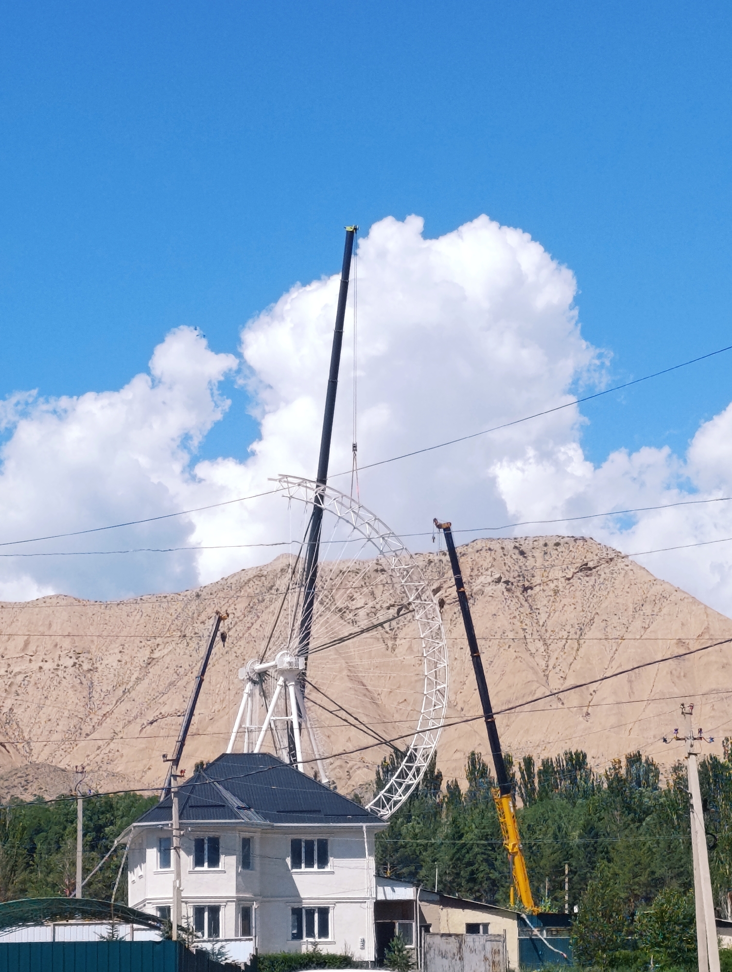 The image depicts a construction scene in Naryn City, Kyrgyzstan, on a bright, sunny day with a clear blue sky and fluffy white clouds. A large Ferris wheel structure, still under construction, dominates the center of the frame. Its white metal framework is partially assembled, with some sections appearing incomplete.
Several construction cranes are actively involved in the assembly. A tall black crane is positioned to the left of the Ferris wheel, its boom extending upwards towards the sky. Another black crane is located behind the Ferris wheel. A yellow crane is visible to the right, its arm angled towards the Ferris wheel.
In the foreground, a modern two-story white house with a dark, sloped roof is situated. It has multiple windows with white frames. To the left of the house, a section of a green fence and a small, covered structure with a green roof are visible. To the right of the house, there are some smaller, single-story buildings.
The background is characterized by a large, arid mountain slope with visible textures and variations in color, ranging from light brown to beige. A line of tall, dark green trees forms a dense barrier between the construction site and the mountain. Overhead power lines are strung across the image, crisscrossing the sky. The overall impression is one of ongoing development and construction in a mountainous urban or semi-urban environment.