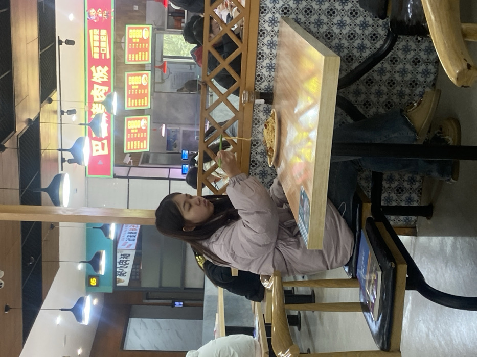 A young woman in a lavender jacket sits at a wooden table in a restaurant. She is using chopsticks to eat noodles from a bowl. Other patrons are visible in the background. Overhead lighting fixtures illuminate the scene, and signage with menu items in Chinese is prominent. The table has a shiny surface and is surrounded by wooden chairs. The floor is tiled with a blue and white pattern.
