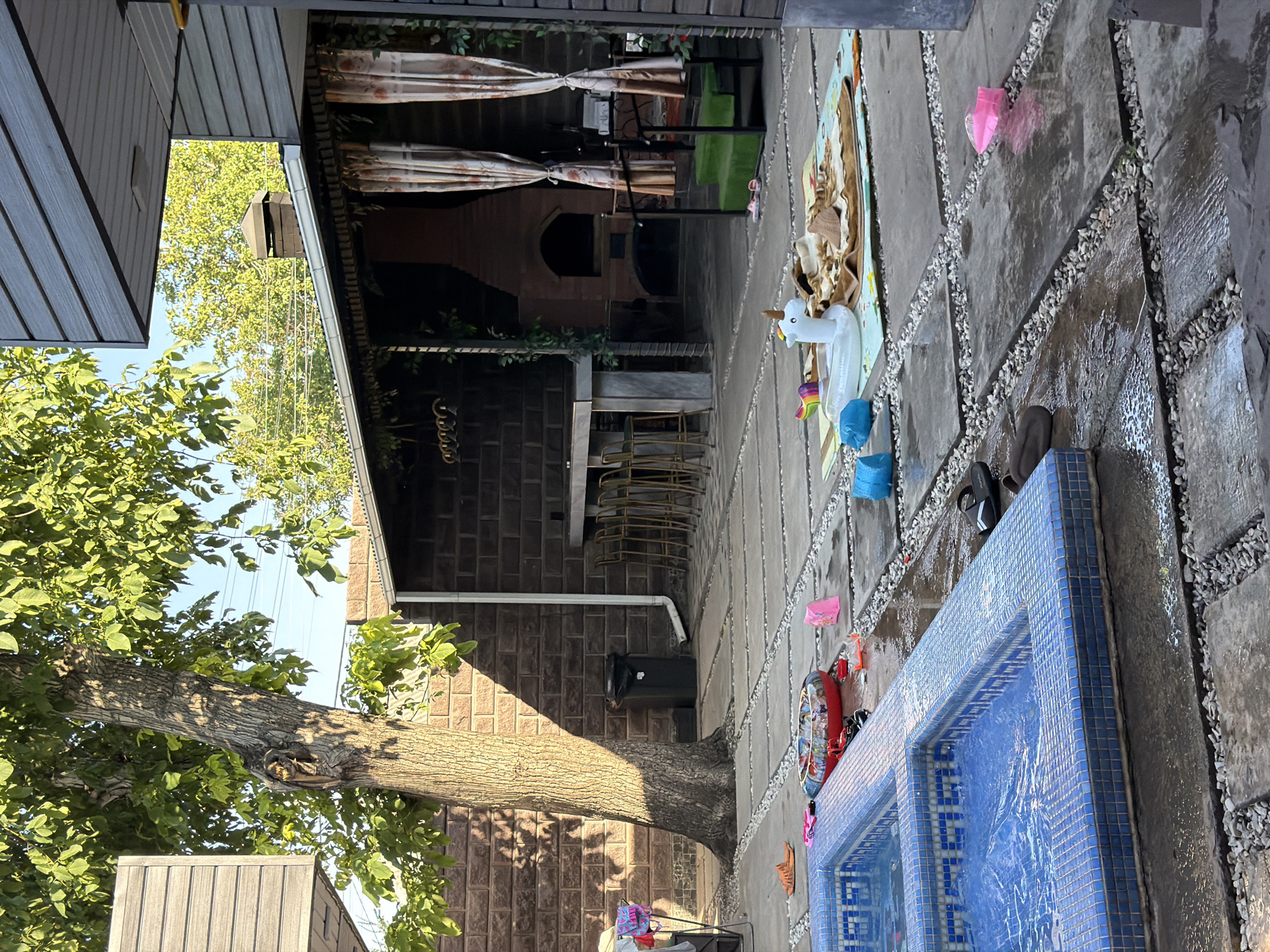 The image shows a backyard patio area in Shymkent, Kazakhstan. A blue tiled pool is in the foreground on the left. The patio is paved with large square stones interspersed with gravel. A large tree with sparse leaves grows on the left side of the patio, casting a shadow. To the right of the tree is a dark trash bin. Beyond the patio is an outdoor covered seating area with a stone fireplace and a long table with bar stools. Curtains hang on the sides of the covered area. Scattered on the patio are various items, including flip-flops, a red inflatable ring, a white inflatable unicorn, a blanket, and several colorful plastic toys. The ground is wet, suggesting recent use or rain. The overall scene depicts a residential outdoor space, likely used for recreation.