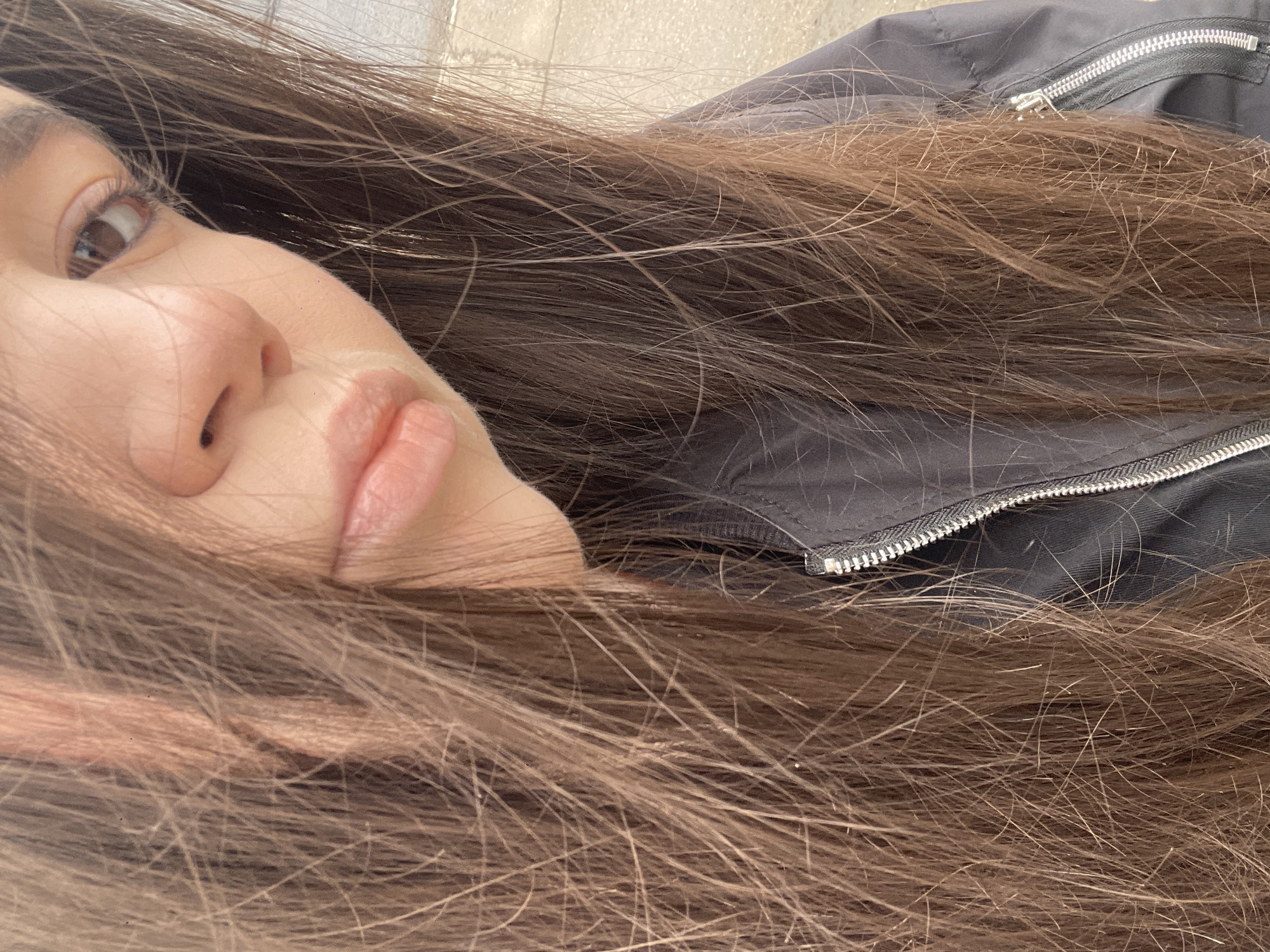 A close-up, canted, indoor shot captures a partial view of a woman's face, predominantly featuring her left eye, nose, and lips. She has fair skin and brown hair that frames her face and is partially covering what appears to be a dark-colored jacket with a visible zipper. The background appears to be a blurred, light, neutral color, possibly an interior wall. The image does not provide sufficient information to determine the location or other details beyond what is immediately visible.