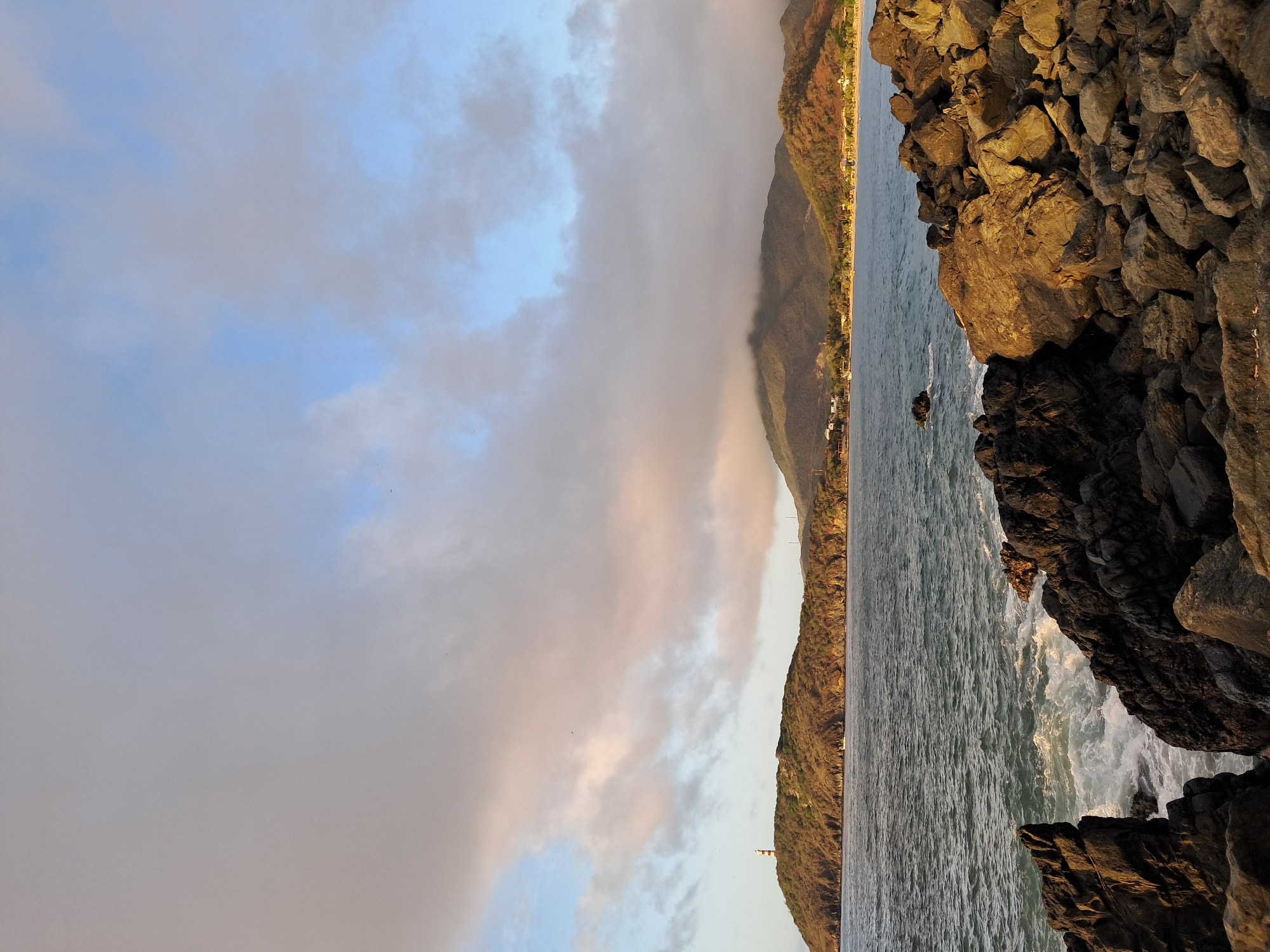 The image depicts a coastal landscape in the vicinity of Pedro González, Venezuela. In the foreground, a rocky shoreline is visible, with dark, jagged rocks in the lower left and lighter, more rounded rocks to the right, illuminated by warm sunlight. Waves are breaking against the rocks, creating white foam. The middle ground features the ocean, with a visible horizon line. In the background, a hilly coastline stretches across the frame. On the leftmost hill, a lighthouse with black and white horizontal stripes stands prominently. Further inland, mountains rise, partially obscured by clouds. The sky above is a mix of light blue and overcast gray clouds, with some areas reflecting the warm light of sunrise or sunset. A few small structures and vegetation are scattered along the coastal hills.