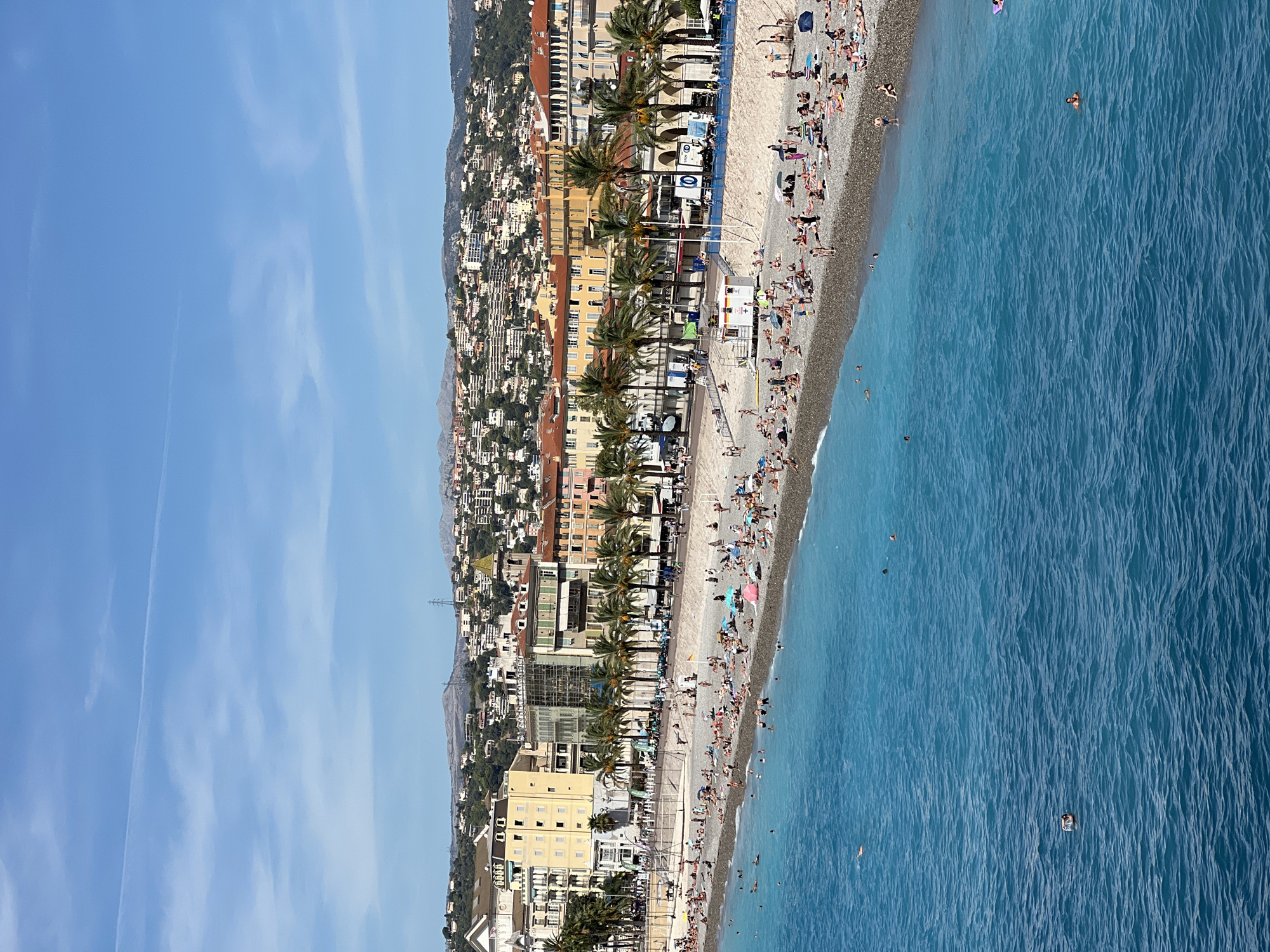 This is a high-angle, wide shot of the Promenade des Anglais in Nice, France, on a bright, sunny day. The foreground is dominated by the vibrant, turquoise waters of the Mediterranean Sea, with gentle ripples and a few scattered swimmers. In the mid-ground, a crowded pebble beach stretches along the coastline, populated by numerous individuals sunbathing, sitting, and wading in the shallows. Beach umbrellas and towels are visible, indicating leisure activities. Beyond the beach, a paved promenade runs parallel to the sea, lined with palm trees and people walking. A row of colorful buildings, characteristic of Nice's architecture, forms the backdrop, with pastel-colored facades and red-tiled roofs. Further inland, a densely built hillside is covered with more buildings and vegetation, leading up to a hazy mountain range under a clear blue sky with thin, wispy clouds. A lifeguard station with a red roof is present on the beach, along with various kiosks and signage. The scene depicts a bustling summer day at a popular tourist destination.