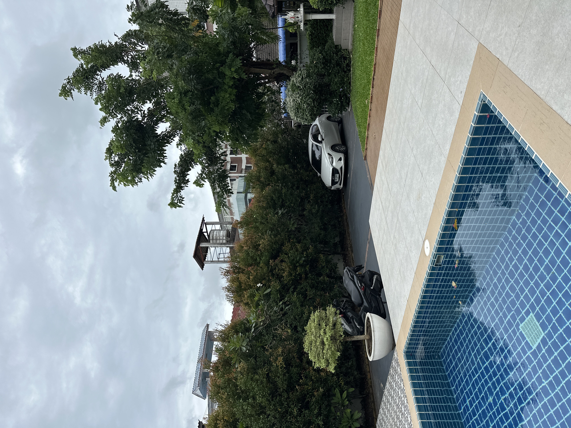 The image depicts an outdoor scene with a swimming pool in the foreground, tiled in blue mosaic. Adjacent to the pool is a paved area with light gray square tiles. Beyond the paved area, a motorcycle is parked next to a potted tree. Further back, a white sedan is parked in a driveway, with a dense hedge lining the background. Above the hedge, buildings and structures, including what appears to be a water tower, are visible against an overcast sky. The environment suggests a residential or commercial property in an urban setting.