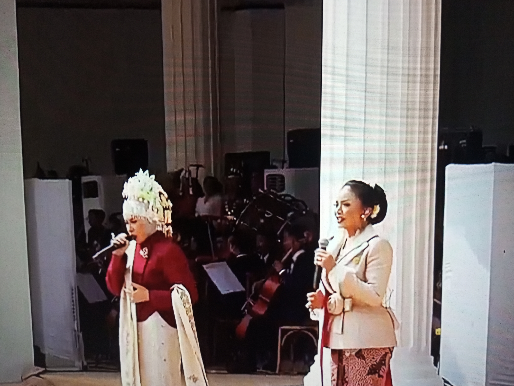 Two women are singing into microphones on a stage. The woman on the left wears a red top and a tall, ornate headdress. The woman on the right wears a light-colored blazer over a patterned skirt. Behind them, an orchestra with cellos and other instruments is visible. The stage is flanked by large white columns and is dimly lit. The image appears to have been taken at an indoor event, possibly a cultural performance or ceremony in Banyudono, Indonesia, given the traditional attire.