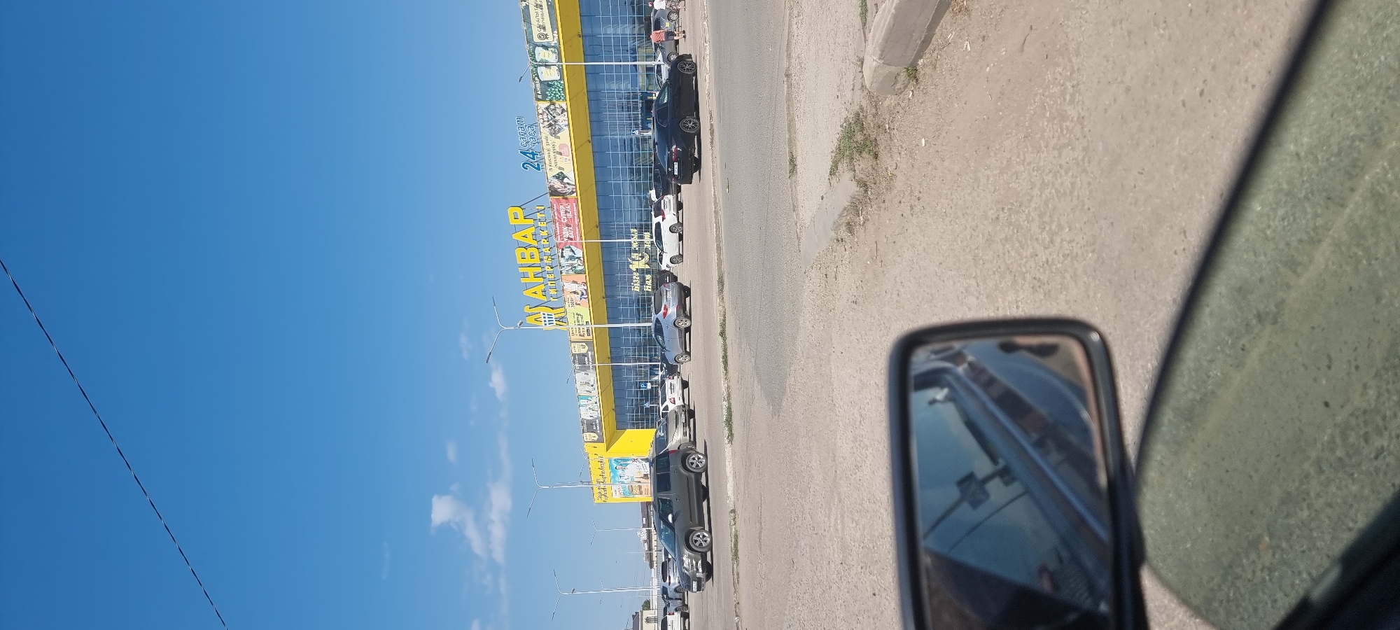 This is a daytime, eye-level, exterior shot of a shopping center and its parking lot. The shopping center has a bright yellow facade with the text "АНВАР" in large yellow letters at the top, and "СУПЕРМАРКЕТ" below it in smaller blue letters. The building features large, reflective blue windows and a banner displaying various images. The parking lot in front of the building is filled with several parked cars of various colors and makes, including a dark sedan, a silver SUV, and a white hatchback. In the foreground, the side-view mirror of a vehicle is visible, reflecting the street and some of the parked cars. The sky is clear blue with a few white clouds. The scene appears to be in a city, likely Aktobe, Kazakhstan, given the Cyrillic text on the building.