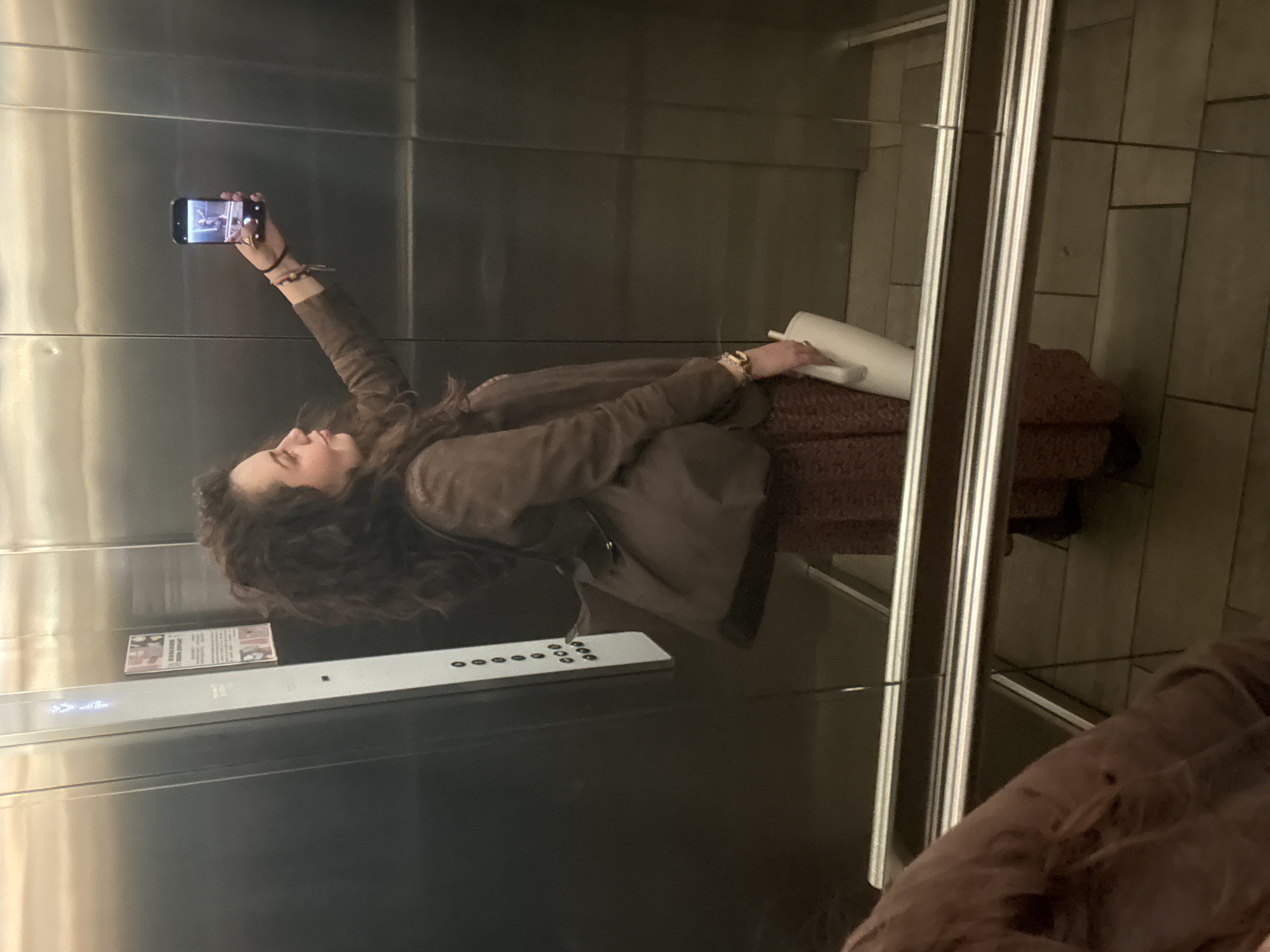 A woman with curly hair is taking a selfie in an elevator. She is holding a smartphone in her right hand, arm extended. She is wearing a brown jacket and a patterned skirt. There is a sign visible on the elevator wall. The location is inferred as Mexico City, Mexico, based on the presence of a sign with Spanish text. A brown coat or jacket is partially visible in the reflection to the right.