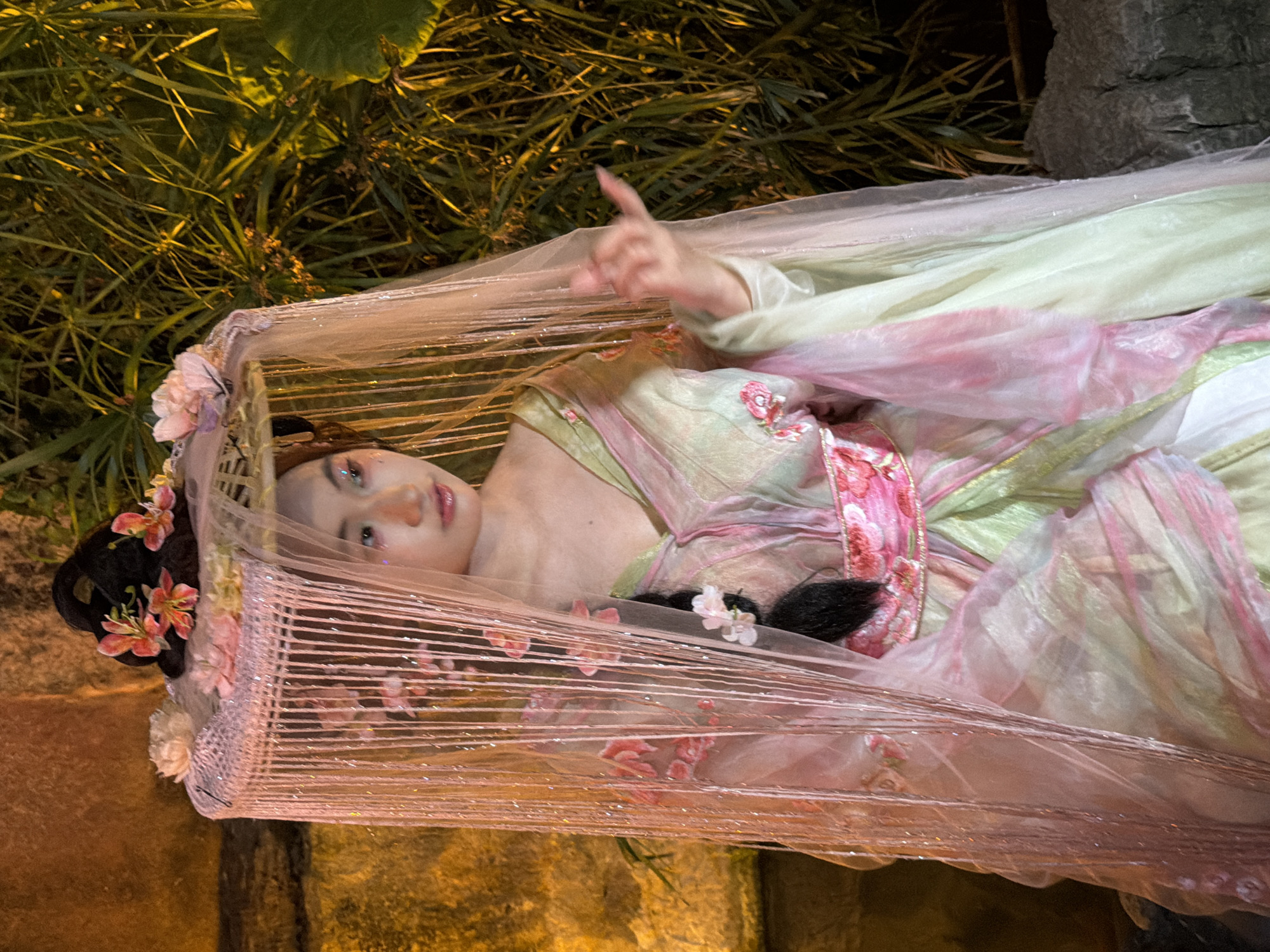 A person, likely a woman, is lying down, possibly on a bed of green foliage. They are adorned in a flowing, traditional Chinese-style gown in shades of green and pink. The garment features intricate floral patterns and delicate, sheer layers that drape over the body. A light, shimmering fabric is draped over them, creating a ethereal effect. The person's hair is styled with flowers and dark accents. Their face is visible, and they appear to be looking upwards with a gentle expression. The scene is illuminated by artificial lighting, creating a dramatic and perhaps mystical atmosphere. The background consists of lush green plants and a rocky surface, suggesting an outdoor setting, possibly a garden or natural landscape within Yuzhong District, China. The overall presentation evokes a sense of beauty and tranquility, with a touch of fantasy.