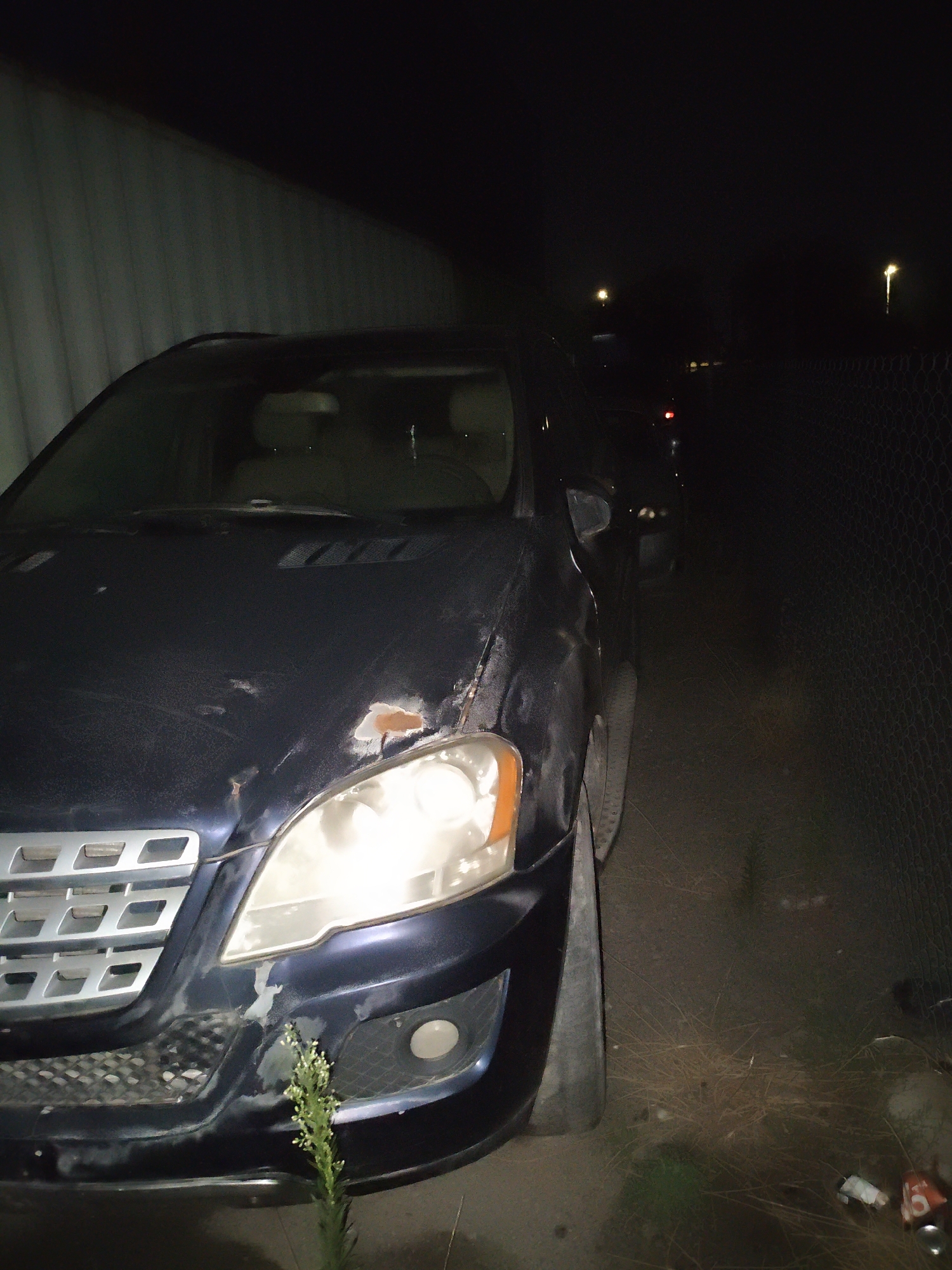 A dark-colored SUV is parked at night next to a corrugated metal wall on the left and a chain-link fence on the right. The SUV's headlights are on, illuminating the front of the vehicle and the ground. The vehicle appears to have some damage, with rust visible on the hood and bumper. The area around the vehicle is unkempt, with dry grass and debris on the ground. In the background, there are streetlights visible, suggesting the location is within a city. Based on the provided information, this scene is located in Krtsanisi, Georgia.