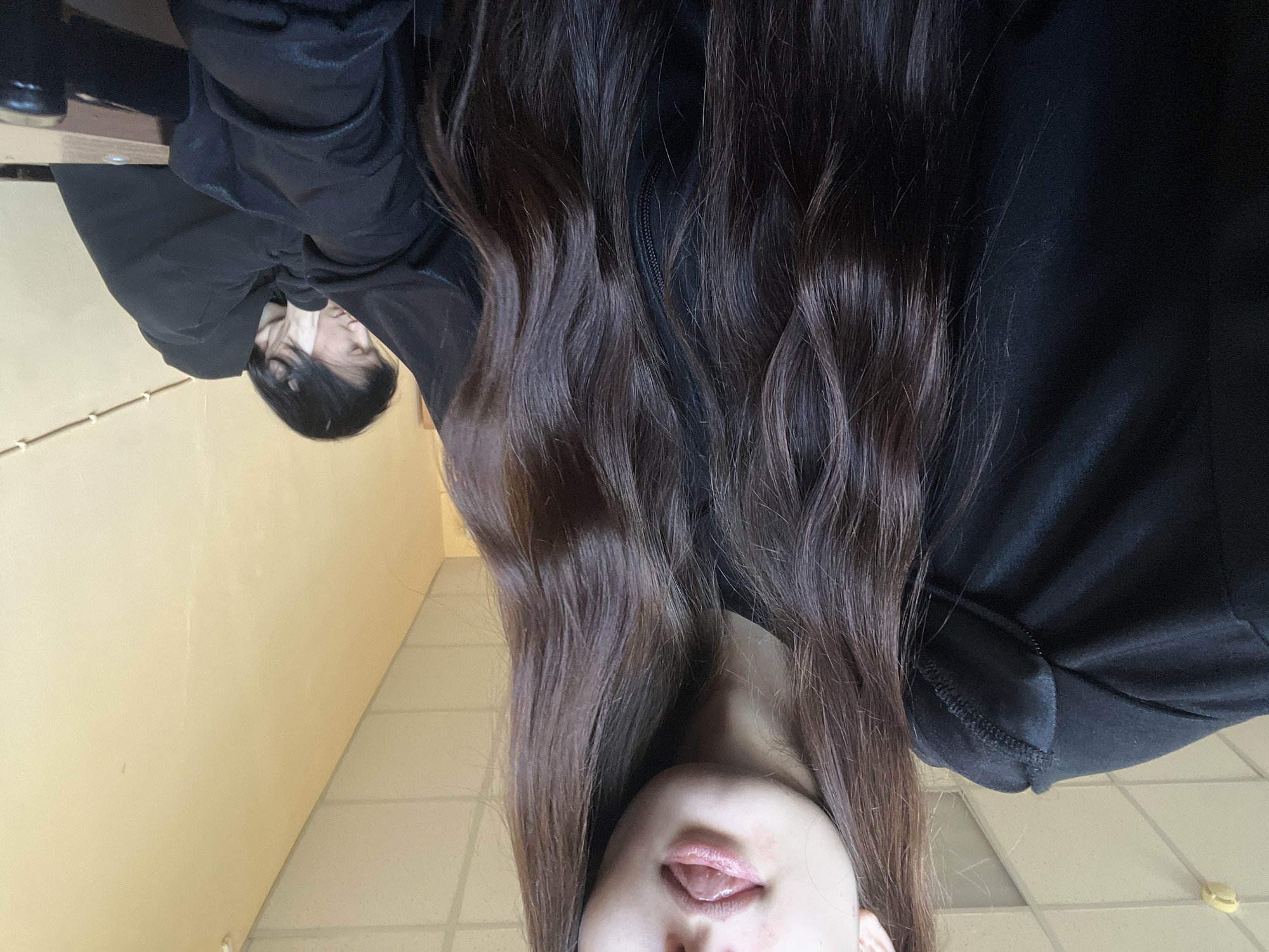 The image displays a close-up, low-angle perspective of a female subject, predominantly featuring her long, wavy, dark brown hair, which frames the bottom portion of her face, revealing her nose and slightly parted lips with her tongue visible. She wears a dark-colored top, possibly a jacket or hoodie, with a zipper detail noticeable amidst her hair on the right side.
In the upper left quadrant of the image, there is a reflection, likely of the same individual or another person, sitting at what appears to be a table or desk. This reflected person has dark, straight hair and is dressed in a dark, long-sleeved garment, with their head resting on their hand, suggesting a moment of contemplation or rest.
The background consists of a plain, light yellow-beige wall that transitions into a light-colored tiled floor. The overall setting appears to be an indoor environment, possibly a classroom, office, or hallway, characterized by functional architecture. The lighting is even and diffused, typical of an indoor space. There is no visible text in the image.