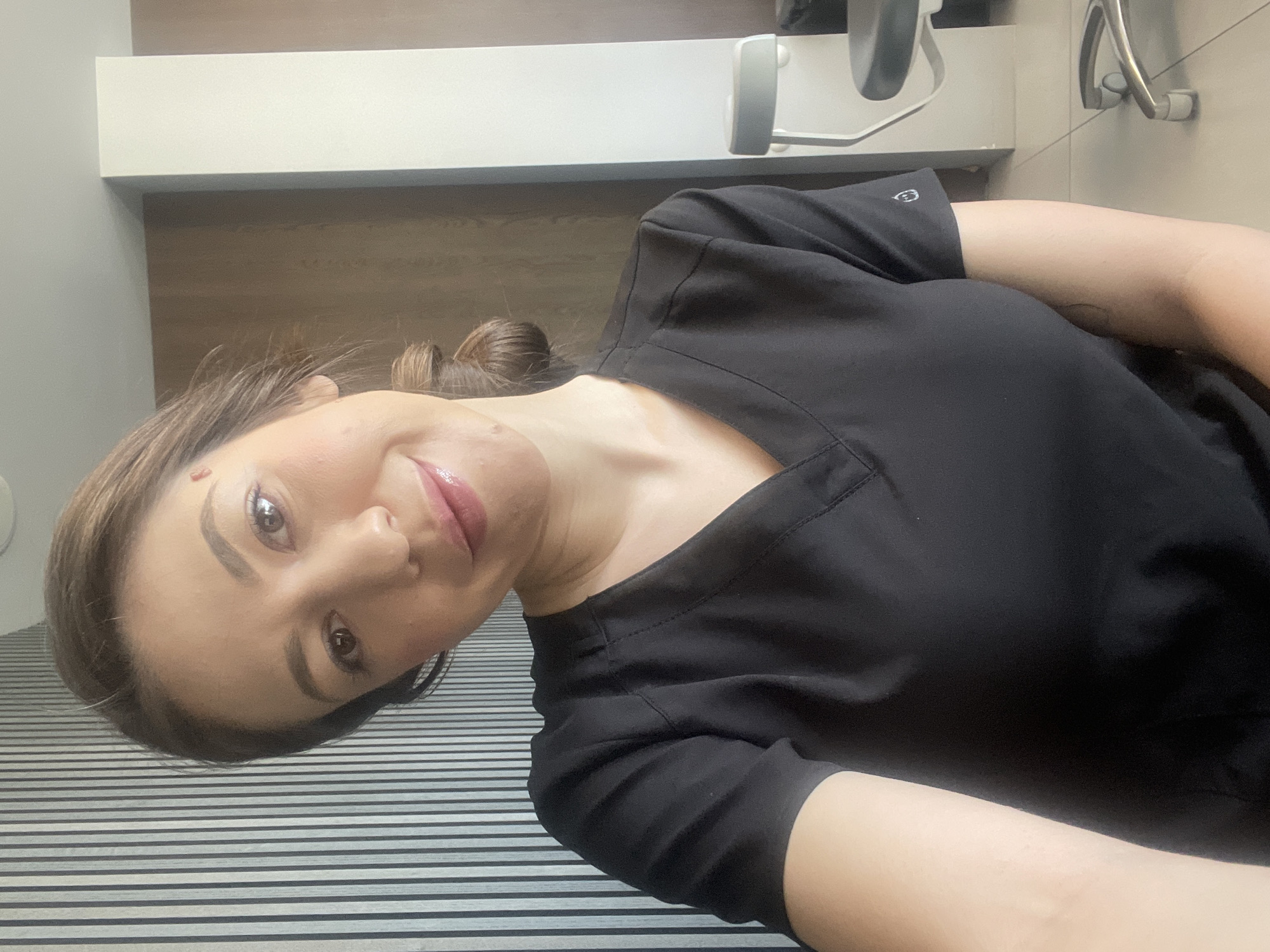 A selfie of a light-skinned woman wearing a black V-neck shirt, likely a medical scrub top, with her hair pulled back in a bun. She has dark eyebrows, dark eyes, and glossy pink lips. The background suggests a medical or dental office setting. Visible in the background are a striped surface, possibly a floor or countertop, and medical equipment (a dental chair) and cabinetry. There is no definitive evidence to specify the location as Sofia, Bulgaria.