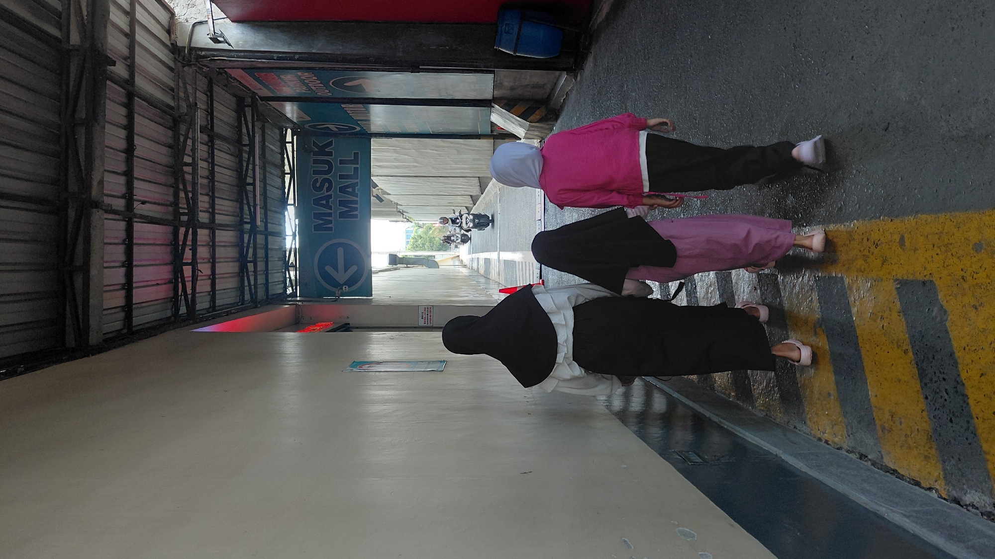 The image shows a narrow outdoor walkway with a covered roof. Three people, two adult females and one child, are walking away from the camera. The person in the middle is a child wearing a purple dress and a black hijab. The person on the left is an adult wearing a white ruffled shirt and a black skirt and hijab. The person on the right is an adult wearing a pink jacket, black pants, and a light blue hijab. Yellow and black hazard stripes are painted on the asphalt ground. A sign overhead reads "MASUK MALL" with an arrow pointing to the left, indicating the entrance to a mall. In the background, two motorcycles are visible, with riders wearing helmets, suggesting this location is in an urban or commercial area. A blue trash bin is visible on the right side of the walkway. The lighting suggests it is daytime, possibly with overcast conditions or shade from the covered roof. The overall scene depicts a group of people entering a mall in Purwokerto, Indonesia.