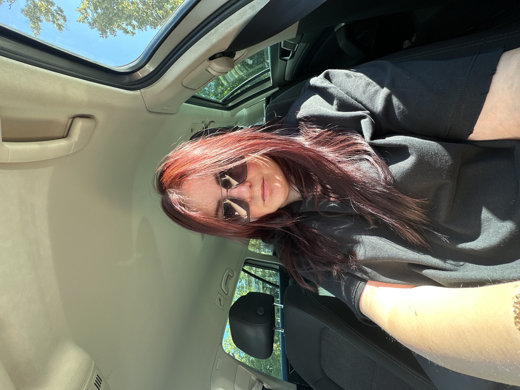 A woman with long, dark red hair and sunglasses is sitting in the driver's seat of a car. She is wearing a black hoodie. The car's sunroof is open, and sunlight is streaming in, casting shadows on her face and clothing. The interior of the car is visible, including the headrest of the passenger seat and part of the dashboard. Outside the car, trees and a clear blue sky can be seen, suggesting an outdoor location. The image is a selfie, taken from the driver's perspective. The scene appears to be in a suburban or rural area, judging by the trees and open sky. The specific location within the city of Zagreb, Croatia cannot be determined from the visual information provided.