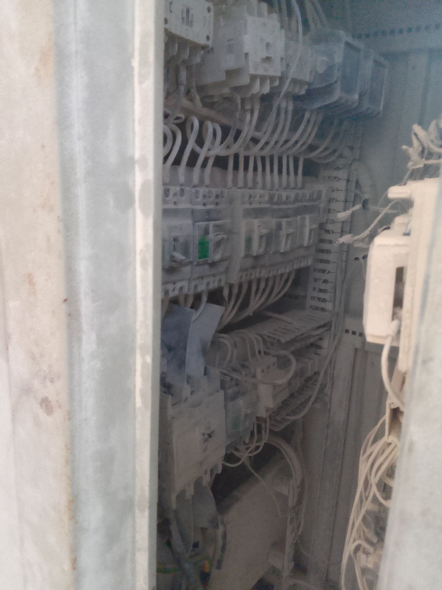 The image shows the interior of an electrical control panel, likely in an industrial setting. The panel is filled with various electrical components, including circuit breakers, relays, and a dense network of wires and cables. The components and wires are heavily coated with a fine, light-colored dust, suggesting an environment with significant particulate matter, possibly from construction or manufacturing. The panel is mounted within a larger, possibly metallic, enclosure. The scene appears to be a close-up view of the complex wiring and electrical infrastructure. There are no people visible in the image.