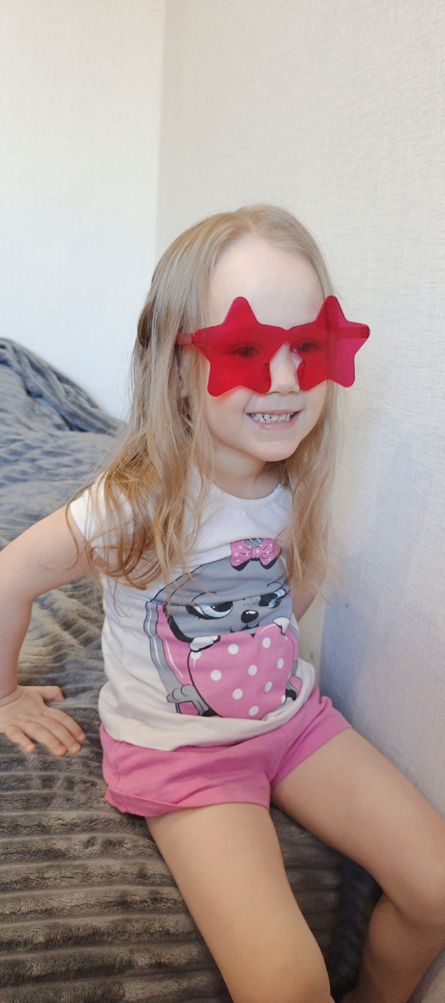 A young girl with blonde hair is seated on a bed, wearing red star-shaped sunglasses, a white t-shirt with a cartoon animal design holding a heart, and pink shorts. She is smiling, revealing her teeth. The background consists of a light-colored wall and a gray textured bedspread. The image appears to be taken indoors. This image was taken in the city of Chaikovsky, Russia.