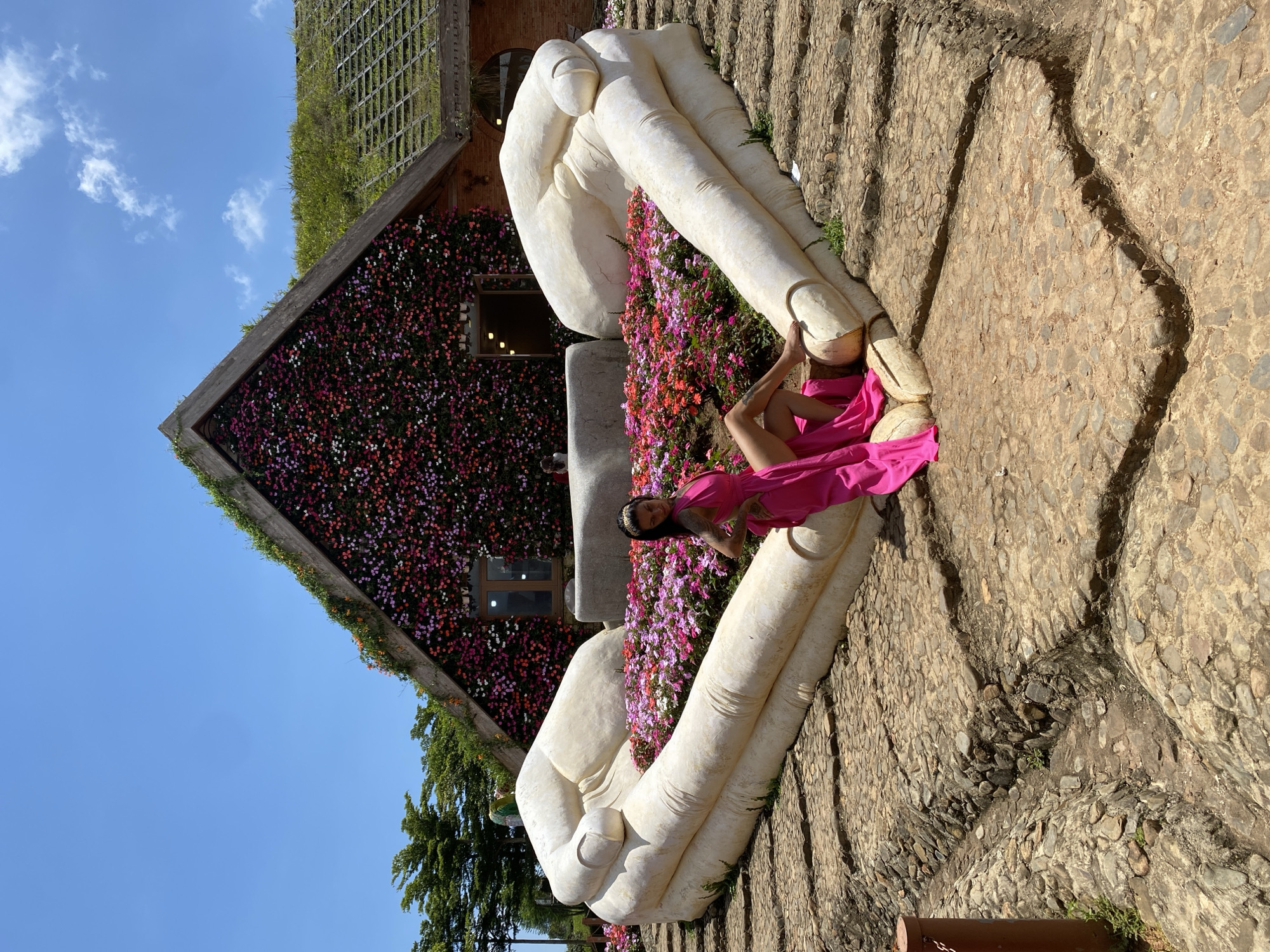 In the city of Cam Ly - Đà Lạt, Vietnam, a woman in a vibrant pink dress poses in a garden. She is seated within a heart-shaped formation created by two large, white, stone hands. Behind the hands, a colorful display of pink and red flowers fills the space. The backdrop is a building with a triangular roof covered in a dense arrangement of flowers, and a green, grassy rooftop. The ground in the foreground is paved with stones, with visible drainage channels. The sky above is a clear, bright blue with a few wispy white clouds. The scene is bathed in natural sunlight, suggesting it is daytime.