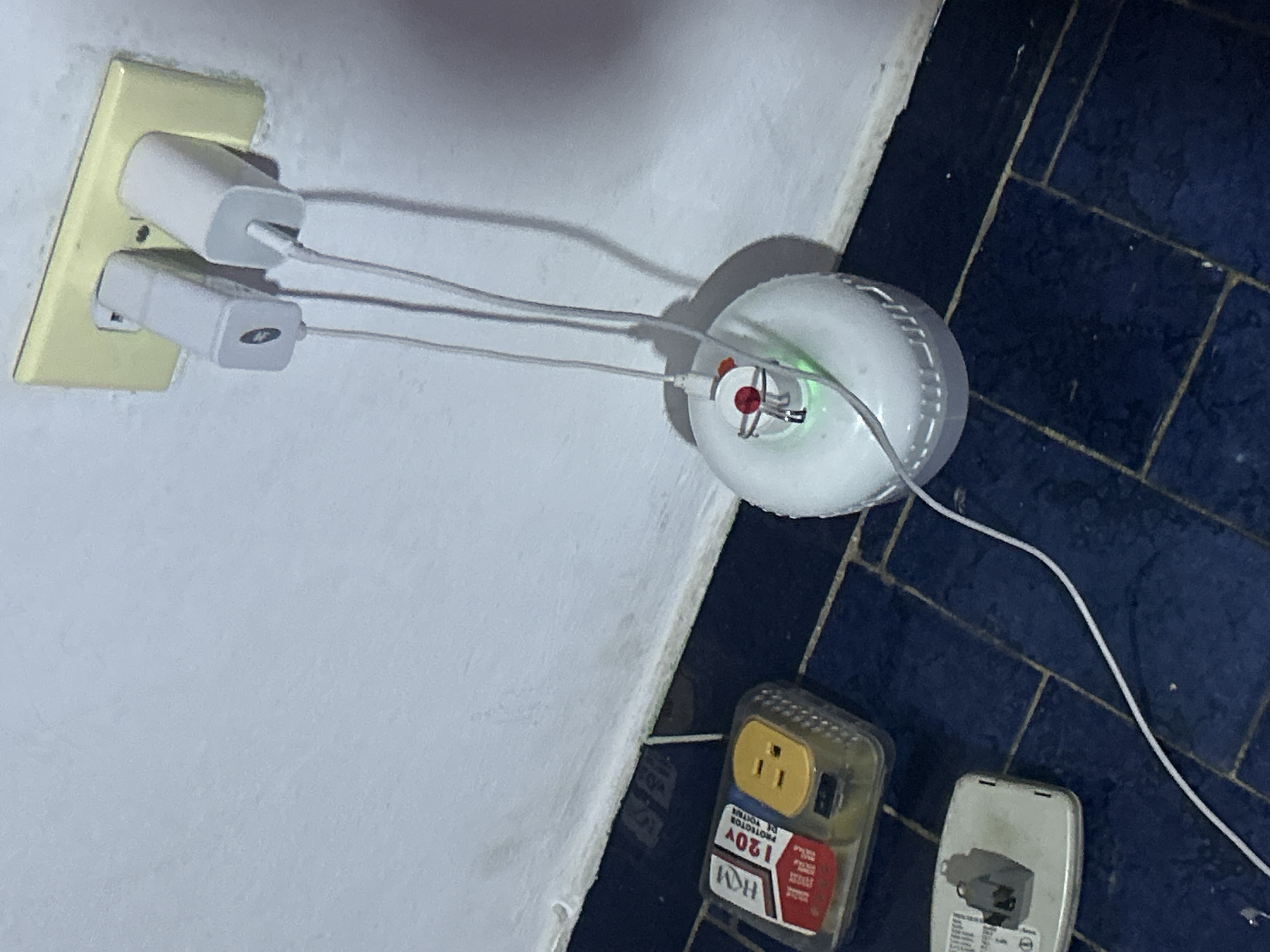 The image depicts a wall with a two-socket electrical outlet occupied by two white chargers. White cables extend from the chargers to a small, white, round device resting on the floor. Next to the round device is a surge protector. The floor is tiled with dark blue tiles. The environment indicates the location is indoors, most likely within a home or office building. The precise location in San Juan de Los Morros, Venezuela cannot be determined from the image. No people or interactions are visible.