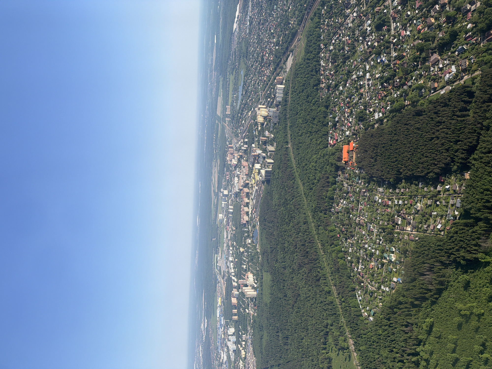 The image shows an aerial view of Перепечино, Russia, with a clear blue sky above. The landscape transitions from a dense forest in the foreground to a mix of residential and urban areas in the distance. Patches of greenery are interspersed throughout the city, and a railway line runs parallel to a large forest. An orange building is visible amidst a cluster of houses in the residential area. The environment appears to be affected by some amount of haze or smog in the distance.