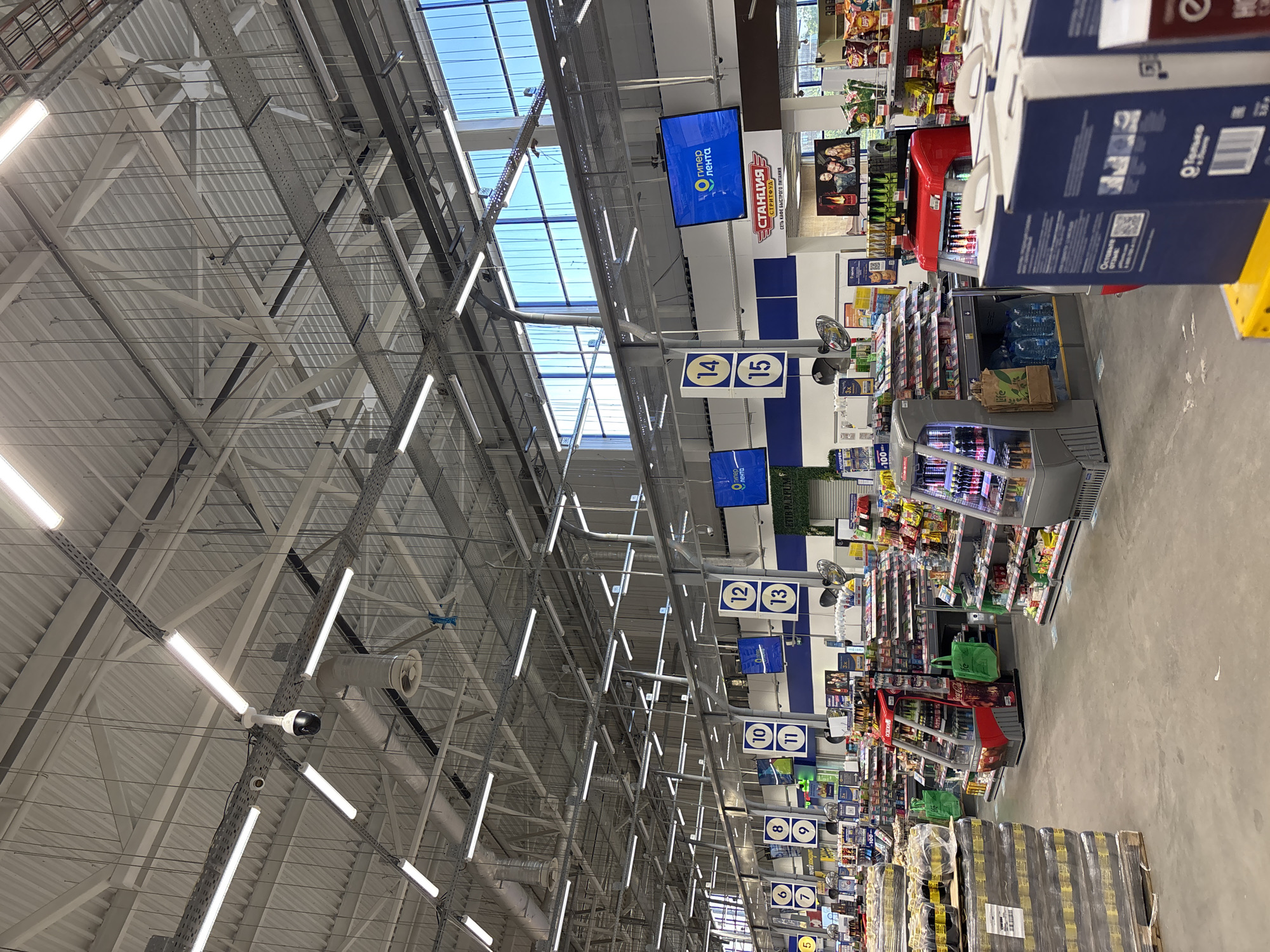 This media file captures the interior of a large hypermarket, likely in Dimitrovgrad, Russia, oriented to show the extensive ceiling structure and multiple shopping aisles. The scene is brightly lit by numerous fluorescent tube lights and natural light filtering through skylights in the high, industrial-style ceiling, which features exposed metal beams, trusses, and ventilation ducts. Surveillance cameras are also visible on the ceiling infrastructure.
The core activity is retail shopping, evidenced by the clearly marked aisles and stocked shelves. Blue and white signs hang from the ceiling, indicating aisle numbers from 6 to 15, guiding customers. Digital displays overhead prominently feature the "гипер лента" (Hyper Lenta) logo. Another significant sign advertises "СТАНЦИЯ СТРИТФУД" (Station Street Food) and "СЕТЬ КАФЕ БЫСТРОГО ПИТАНИЯ" (Network of fast food cafes), suggesting a food service area within the store.
Along the visible aisles, refrigerated display cases contain beverages, and numerous pallets of bottled water are stacked on the floor. Shelves are stocked with various products, including snacks like chips, and other packaged goods. A red shopping cart is partially visible near a display. Text such as "Оставьте отзыв:" (Leave feedback:), "100-", "3x", and "ETIB PARFUM" can be seen on various labels and signs. The floor is a light, polished concrete. The setting is clean, organized, and appears to be bustling with merchandise, though no specific individuals are clearly visible in the frame. The overall impression is that of a modern, well-maintained supermarket environment during daytime hours.
