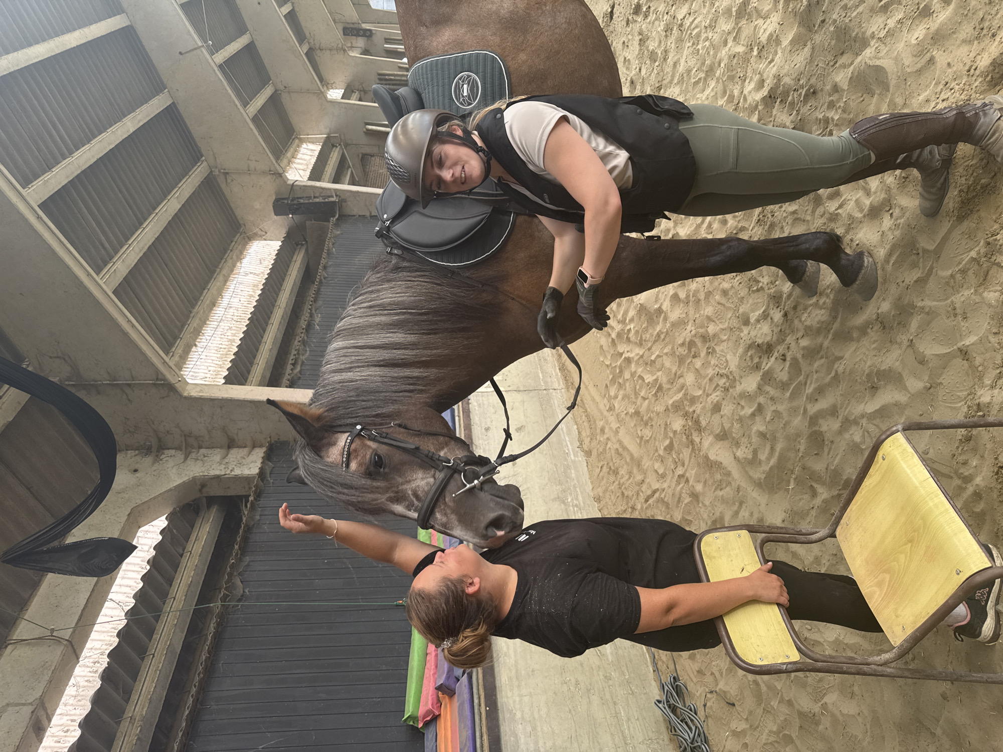 In Estaimpuis, Belgium, two women are interacting with a horse inside a riding arena. The arena has a sand floor and a large, covered roof structure with visible beams and corrugated metal. The walls of the arena are made of concrete and dark paneling.
One woman, wearing a black t-shirt, is positioned to the left of the horse, leaning in to pet its face and appears to be whispering to it. She is also holding a small yellow chair. The second woman, dressed in riding attire including a helmet, vest, and riding pants, is on the right side of the horse, holding its reins and looking towards the camera. She is wearing black gloves and a smartwatch on her left wrist. The horse is a dark brown color with a saddle and bridle. In the background, colorful mats are leaning against a wall, and a black net hangs from the ceiling. The overall scene depicts a moment of connection and preparation within a equestrian setting.