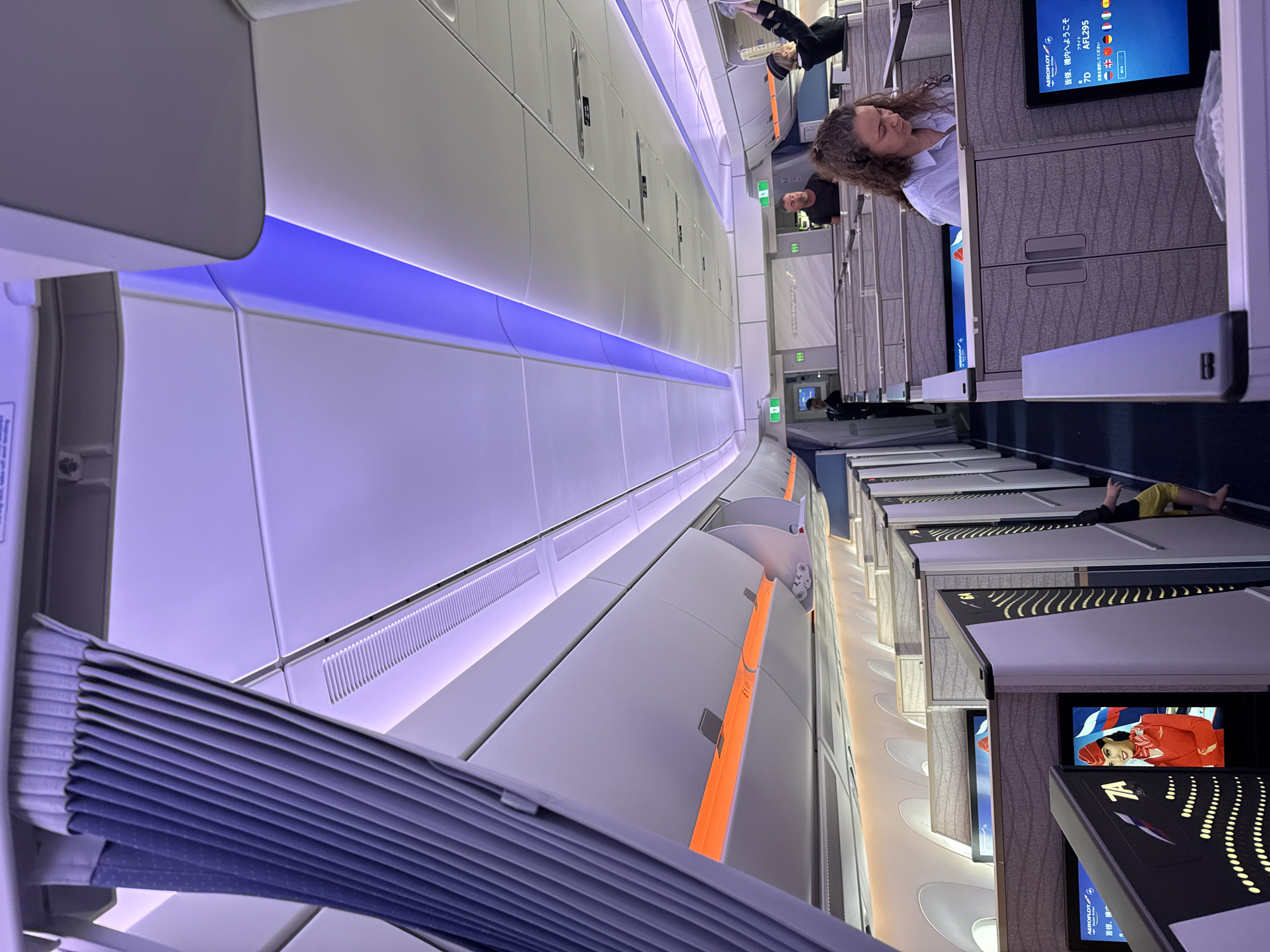 Here is a detailed description of the image:
The image shows the interior of a cabin, possibly an airplane's business class section, with rows of individual suites or seating areas. These suites have privacy partitions and entertainment screens showing various content, including promotional images and flight information in languages like Japanese.
The lighting in the cabin is a combination of soft ambient light, with blue and orange accent lighting strips along the overhead bins, and light from the entertainment screens.
People can be seen both seated and standing in the aisle. One woman is leaning over the suite, looking at something beyond the partition. Another person is reclining in one of the suites, with their feet extending towards the aisle.
The environment suggests a high-end travel setting, emphasizing privacy and comfort.